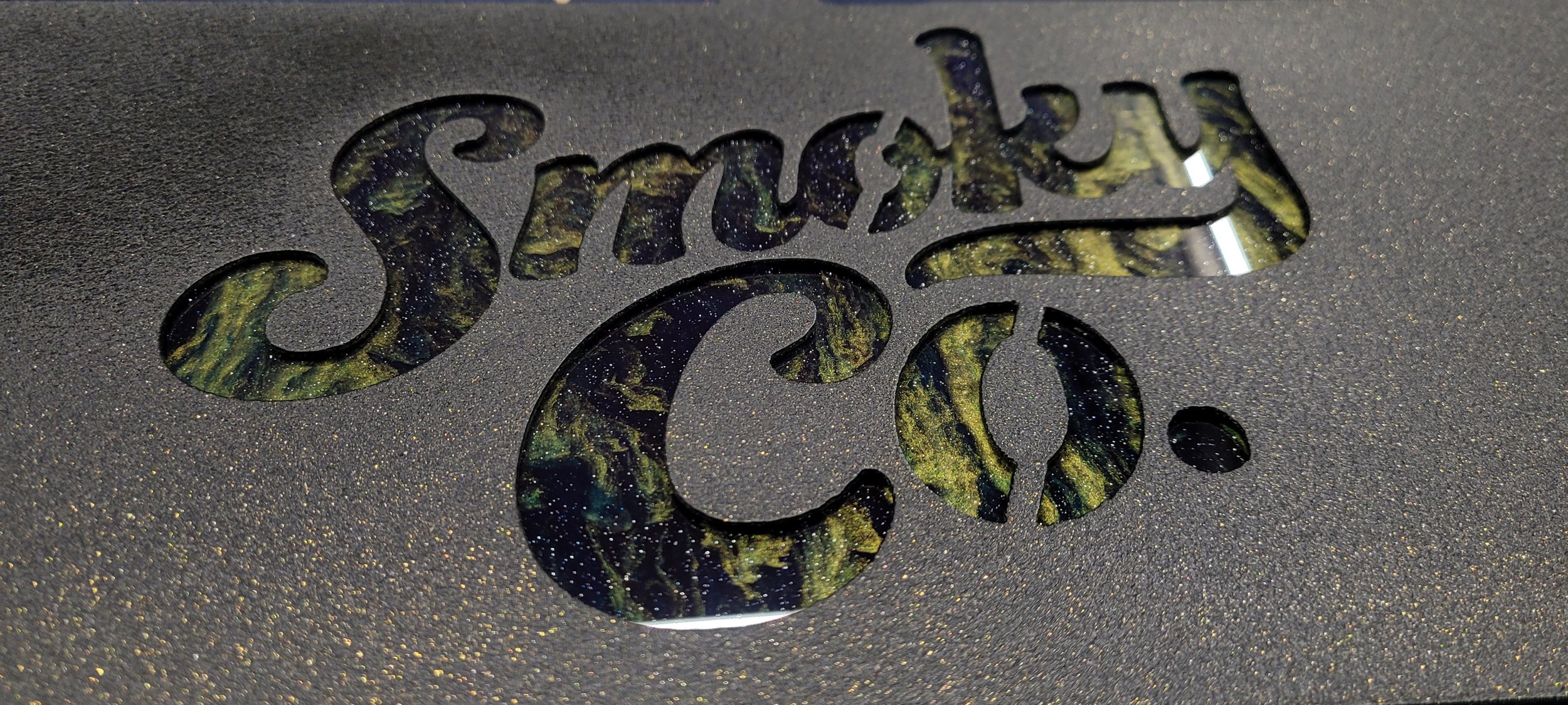 Close-up of a metallic surface with the word 'Smack' cut out, revealing a swirling green, gold, and black pattern underneath. Custom made metal sign