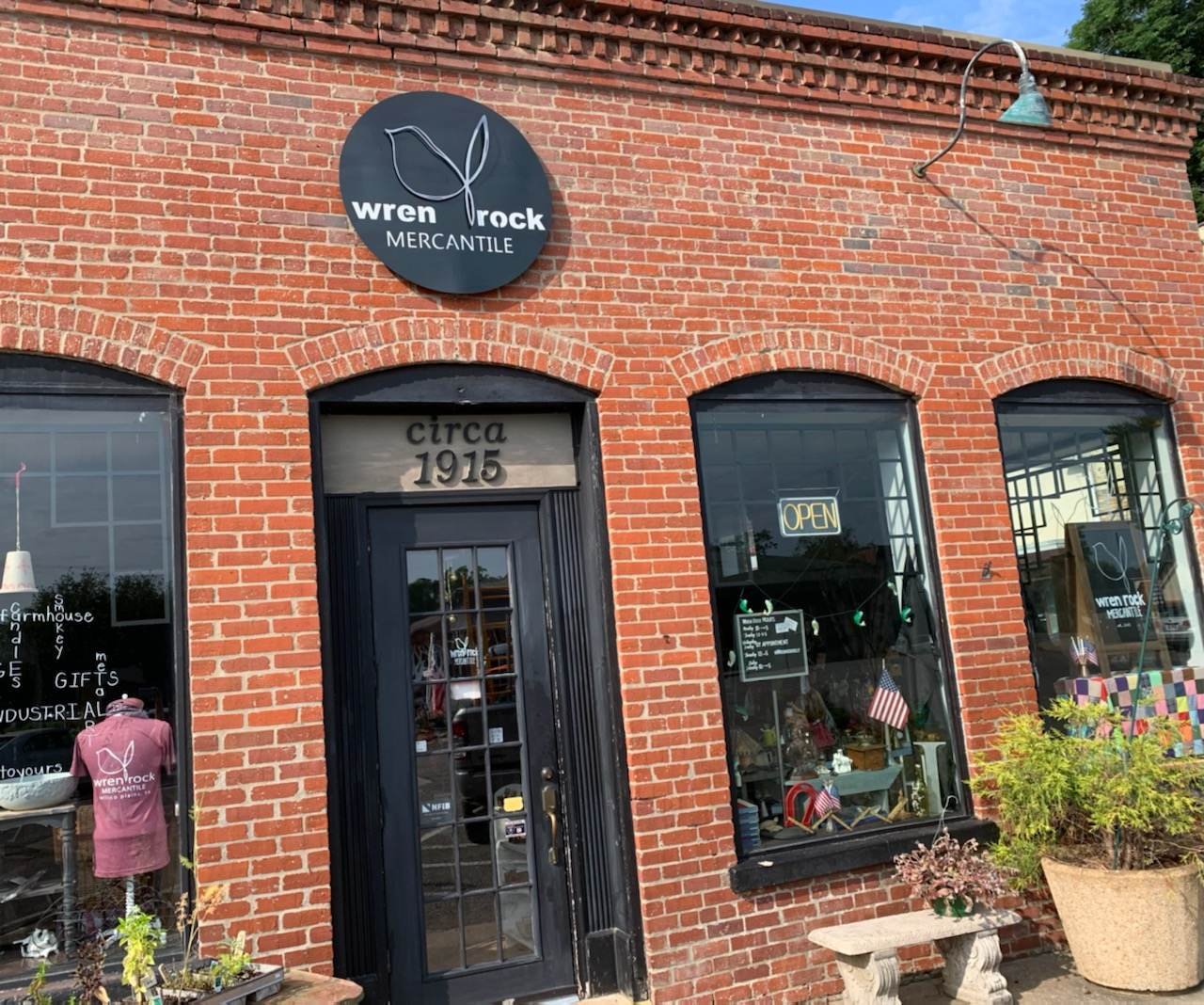 Brick storefront of Wren Rock Mercantile with a round sign featuring a leaf logo, the store name, and opening hours, and large display windows showing products and a neon 'Open' sign.