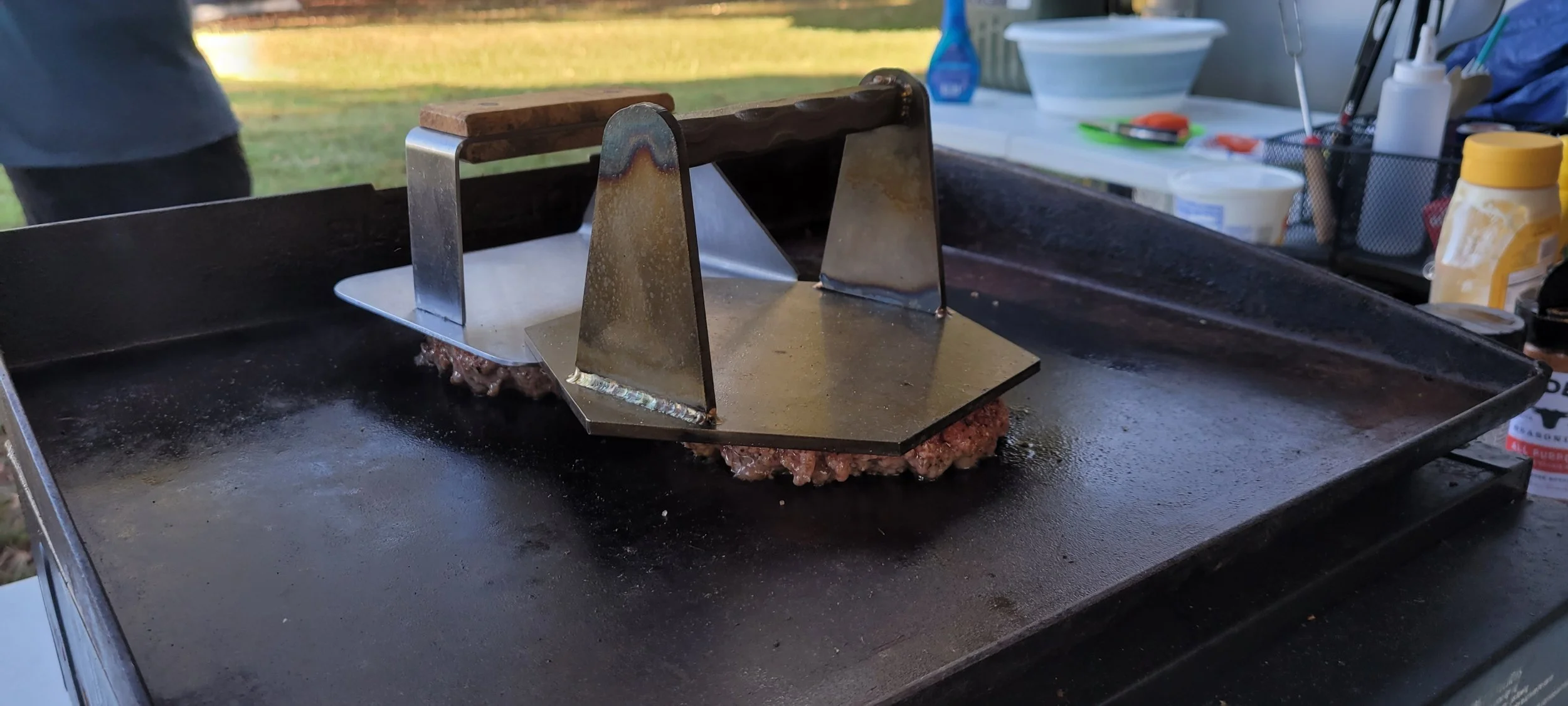 A flat-top griddle with a metal press on ground meat patty, outdoors with kitchen supplies in the background.