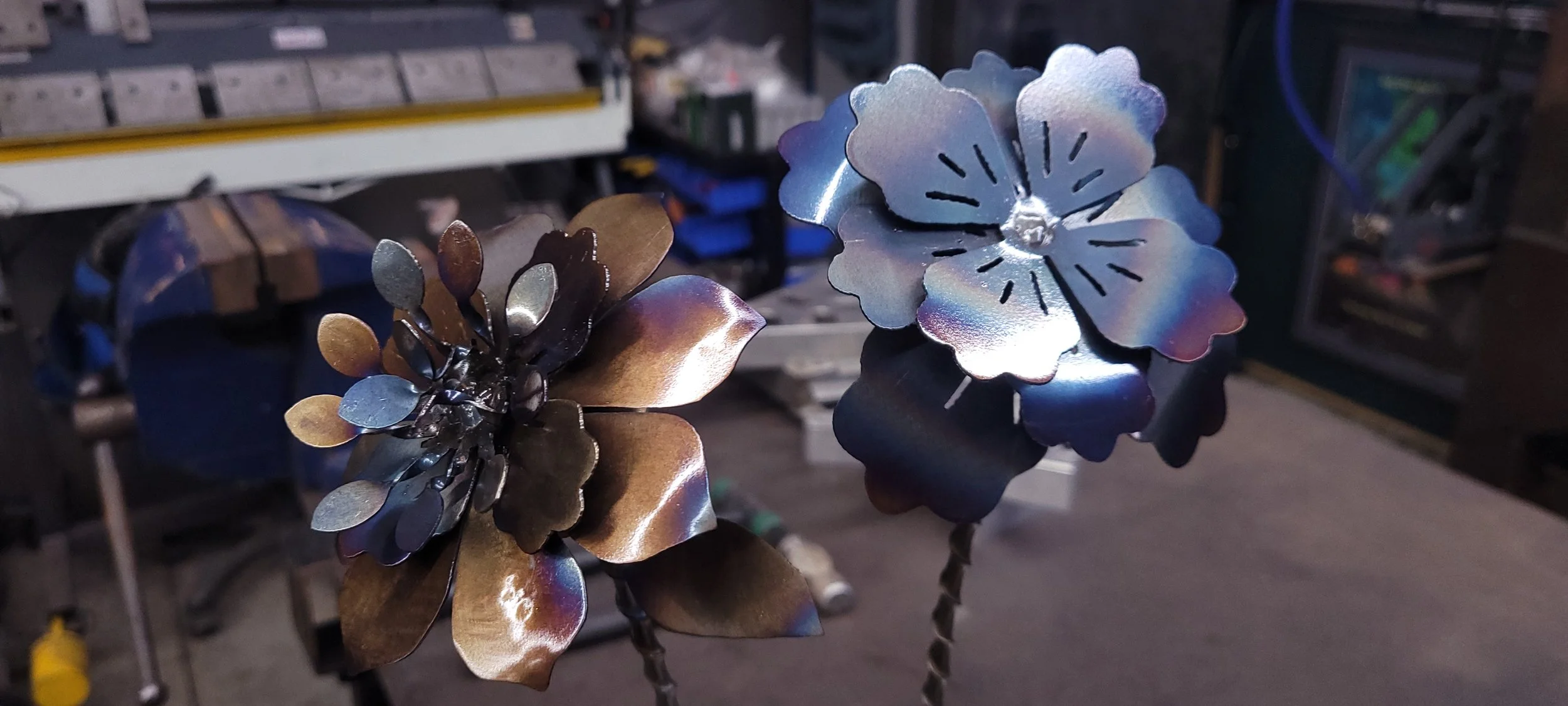 Close-up of metal flowers, some darker, some iridescent, in a workshop environment.  Custom plasma cut metal flowers fabricated by Turn 2 Customz in Tellico Plains Tennessee