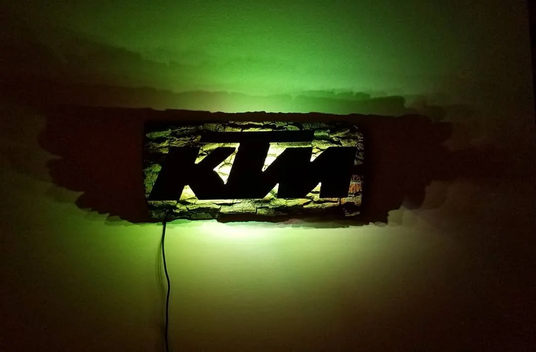 Wall-mounted KTM logo sign with backlighting, illuminating the surrounding wall in green and casting shadows.