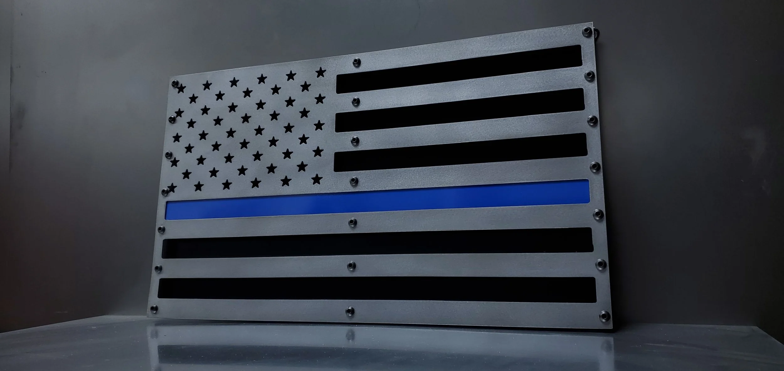 Metal American flag with black and blue stripes, star pattern, and screws, on a metallic gray wall.