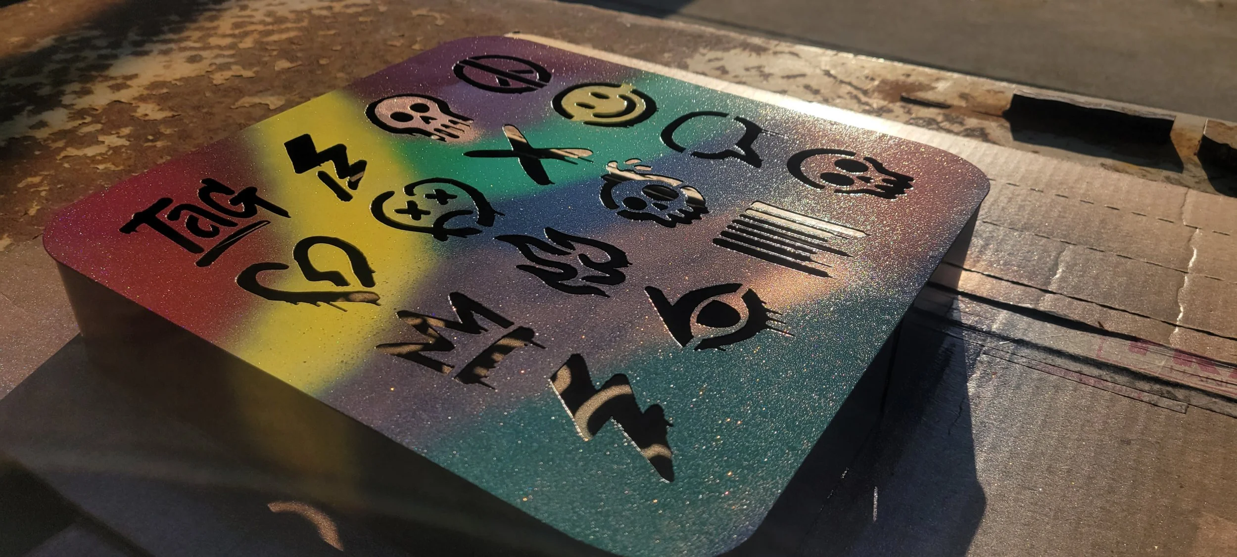 A colorful, glittery stencil art piece on a rectangular metal surface with cut-out symbols and words, placed on a wooden surface. Made by Turn 2 Customz