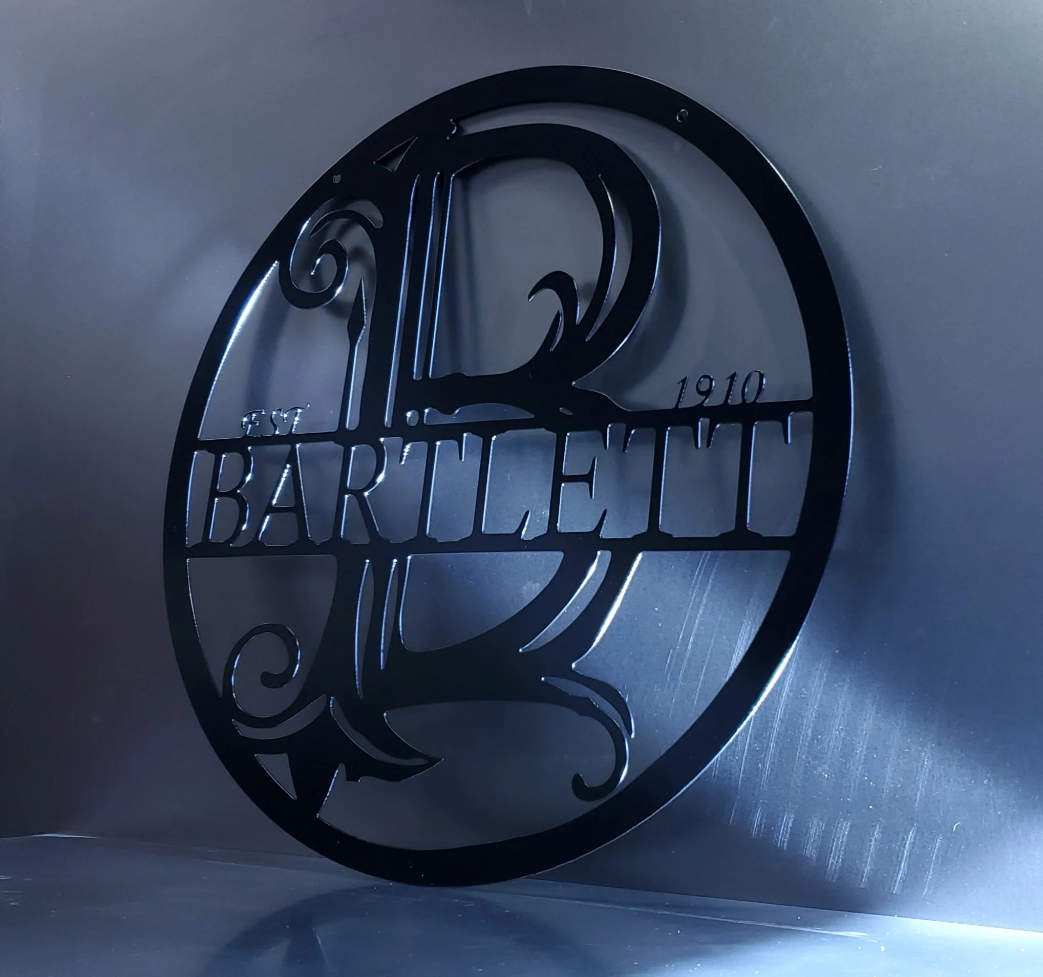 A round black metal sign with the name "Bartlett" and the year "1910" on it, featuring decorative scrollwork and a stylized tree, mounted on a gray wall.
