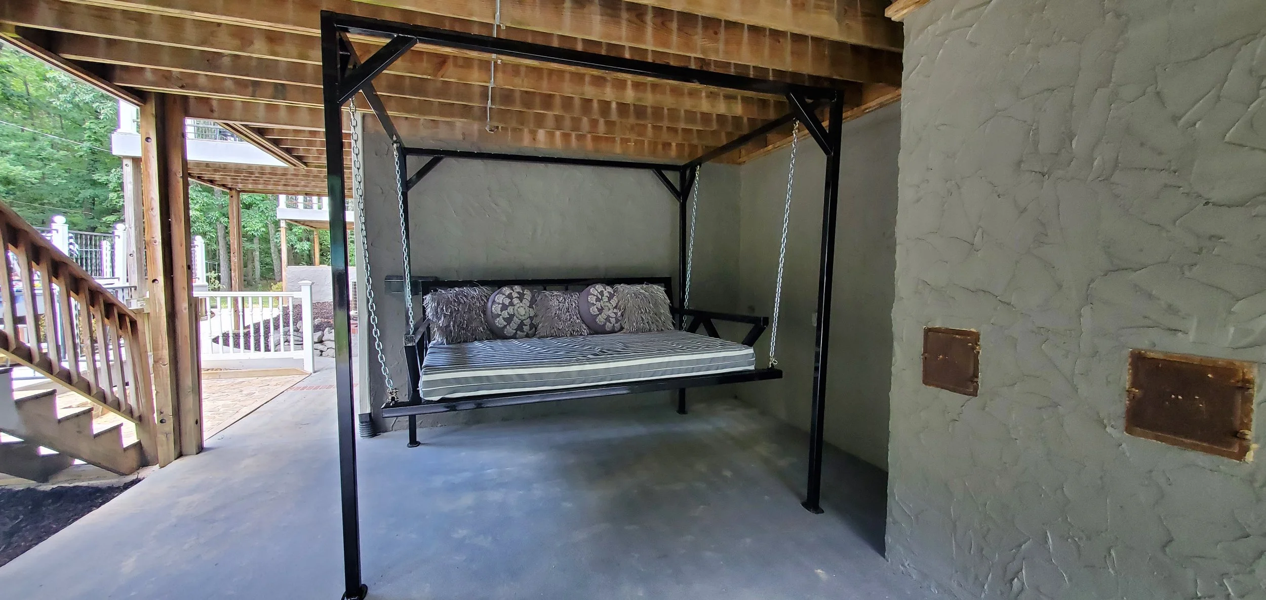 A black outdoor swing bed with a cushion and multiple pillows, hanging from a frame under a wooden deck in a backyard.