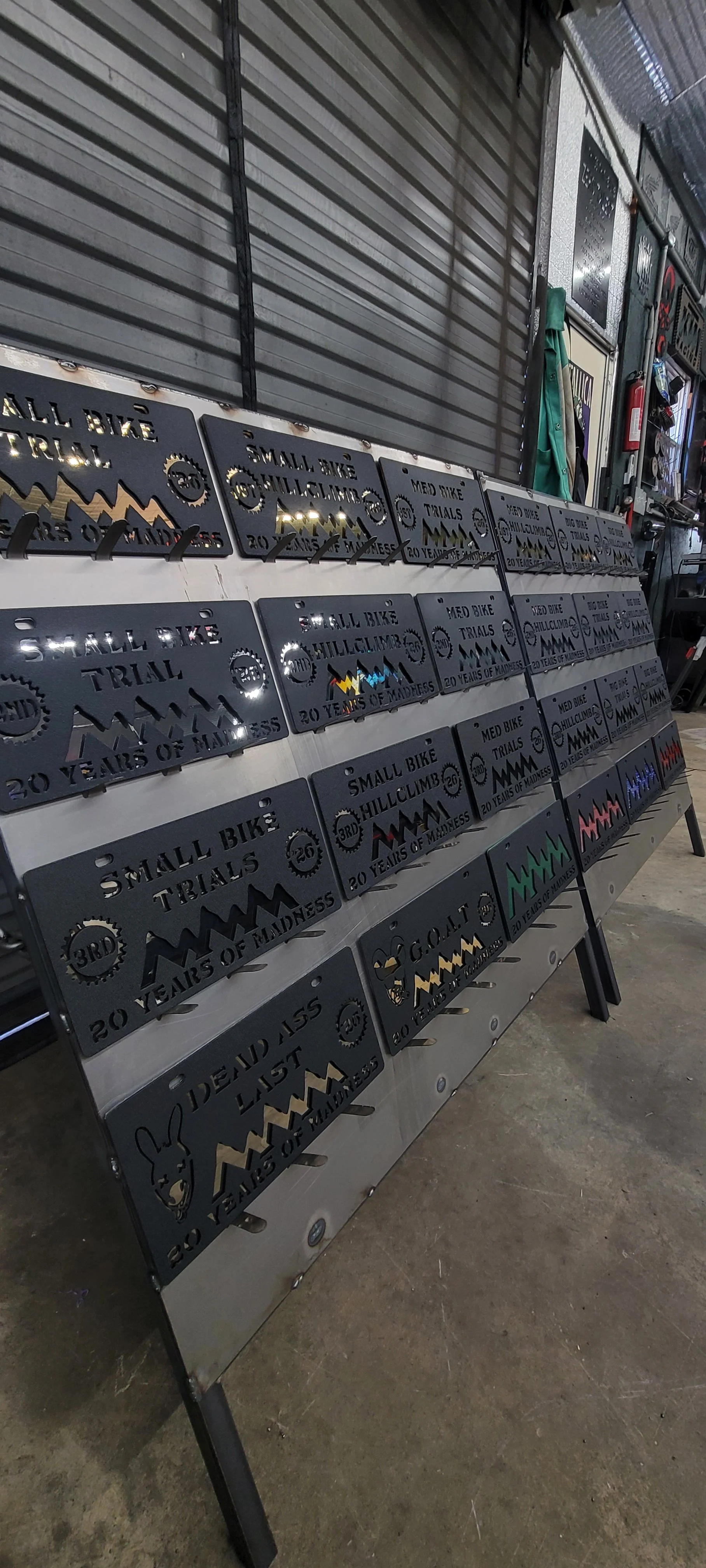 A display of metal plaques with cut-out text and graphics celebrating 20 years of madness for a small bike trial event, arranged on a metal rack inside a garage or workshop. Made by Turn 2 Customz