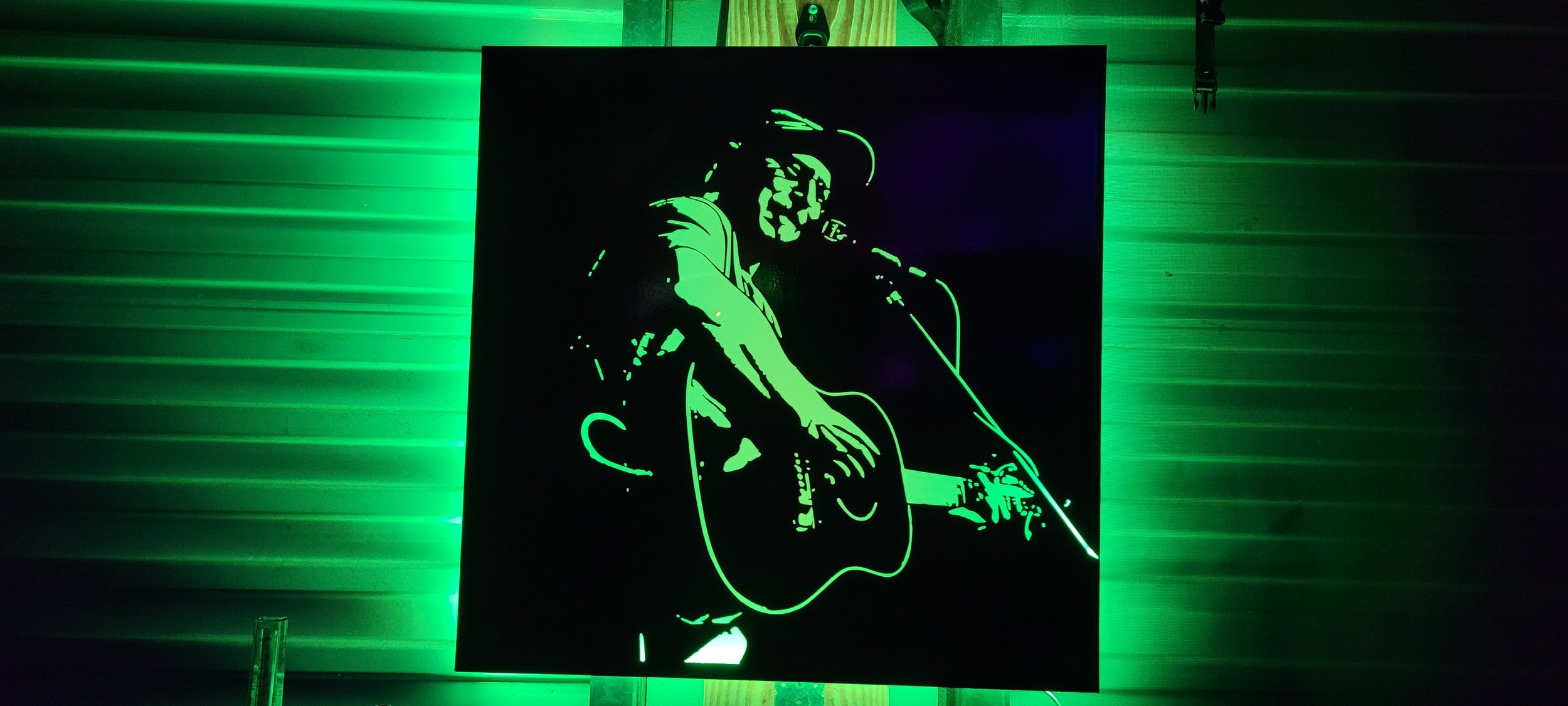 A neon-lit silhouette of a musician playing an electric guitar, with a green glow, mounted on a black background. Made by Turn 2 Customz