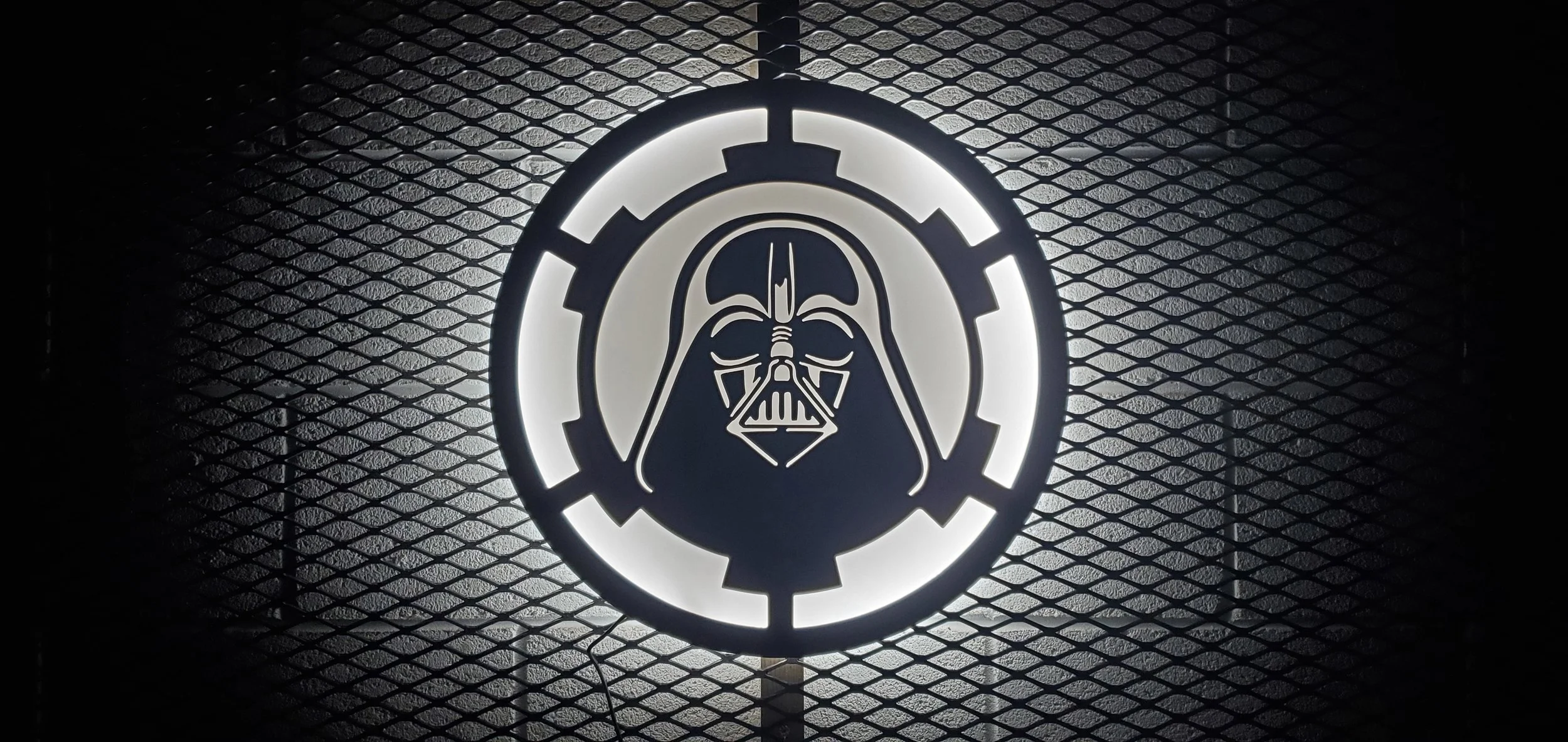 Illuminated sign featuring Darth Vader's helmet from Star Wars inside a circular frame with a gear-like border, mounted on a textured dark background.