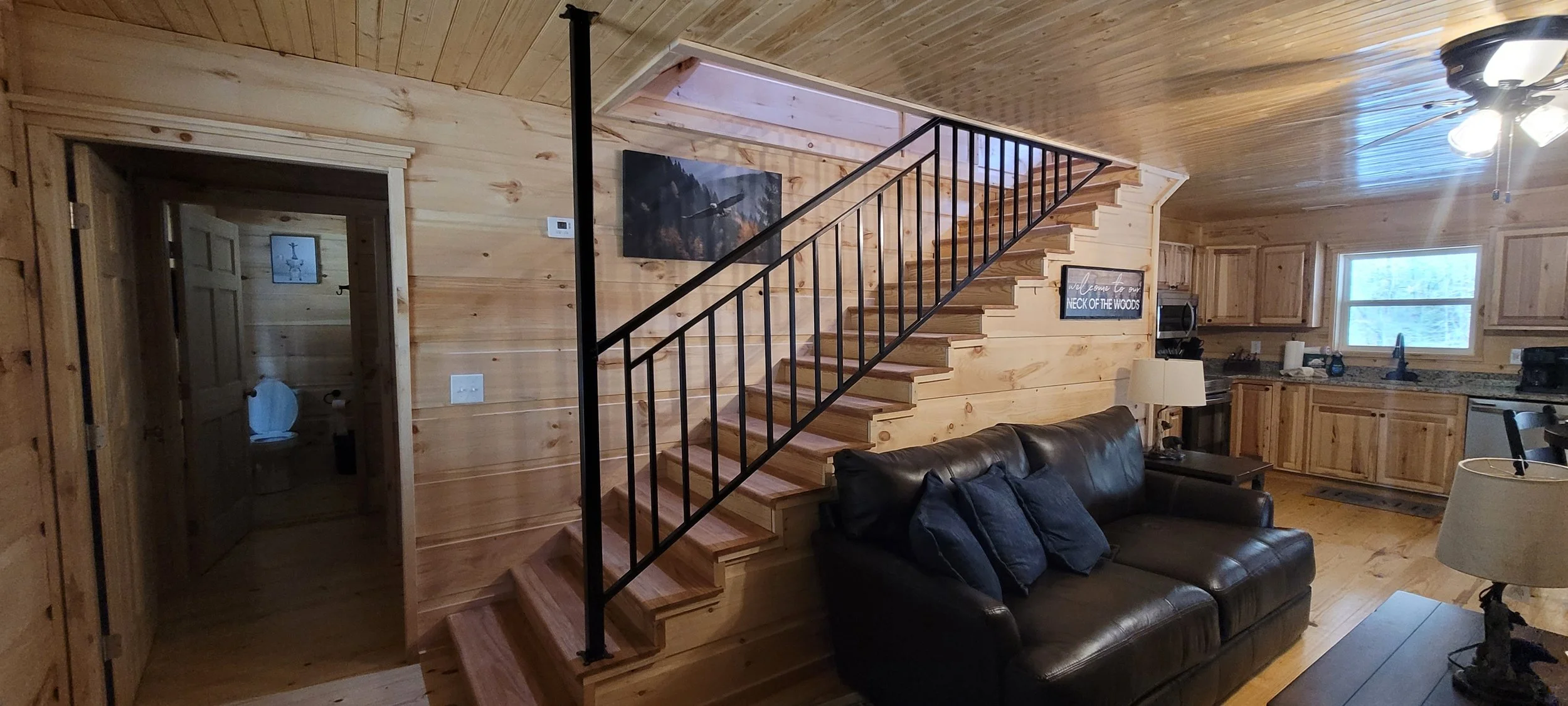 Interior of a cabin living room with wooden walls, a staircase, a leather sofa, and a kitchen in the background.