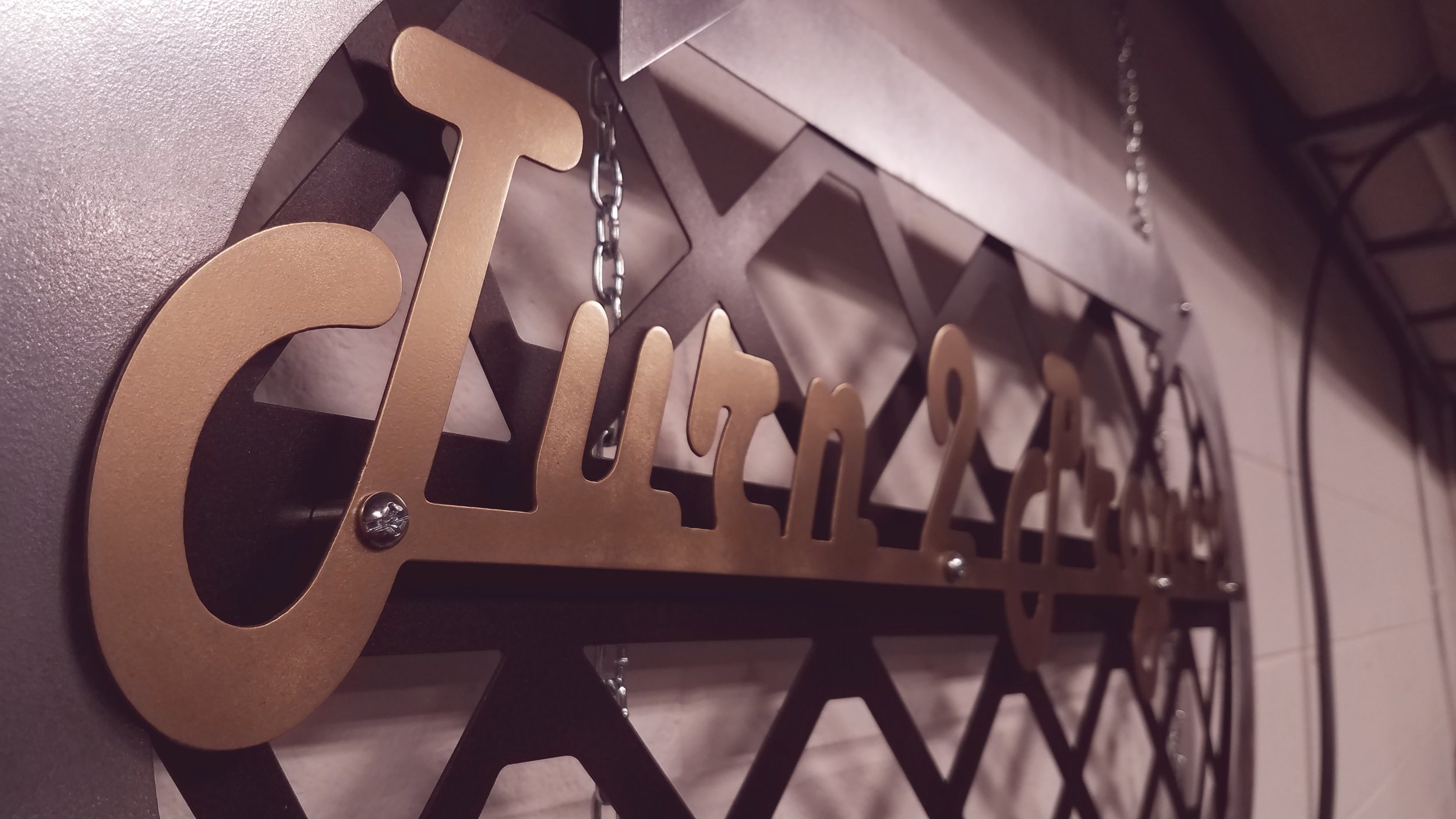 Close-up of a metallic sign with the word 'Tea' hanging from a chain, mounted on a wall. Custom metal sign built by Turn 2 Customz
