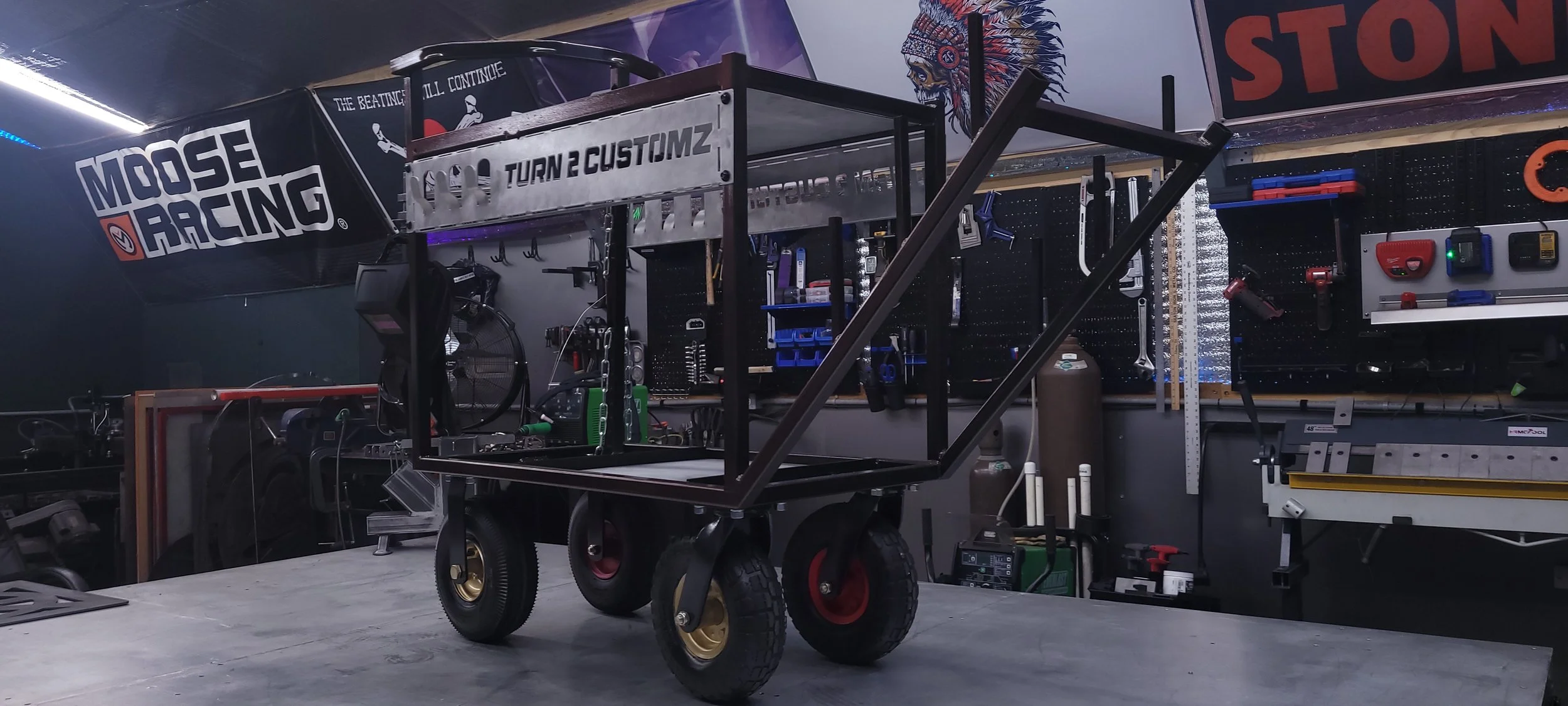 A garage workspace with a partially assembled vehicle frame on a wheeled cart and various tools hanging on a pegboard wall.