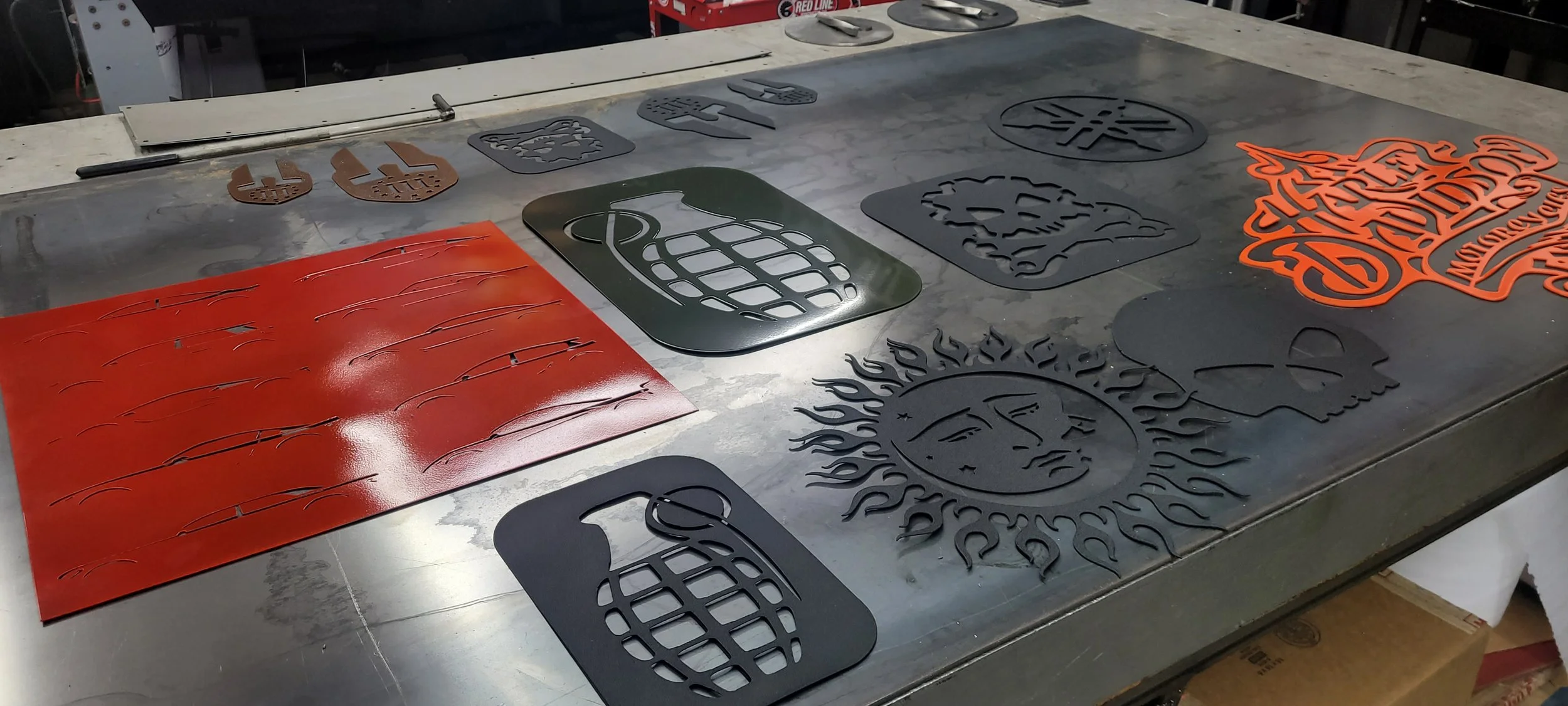 Various metal cutouts and stencil sheets with designs and patterns spread out on a metal worktable in a workshop. Made by Turn 2 Customz