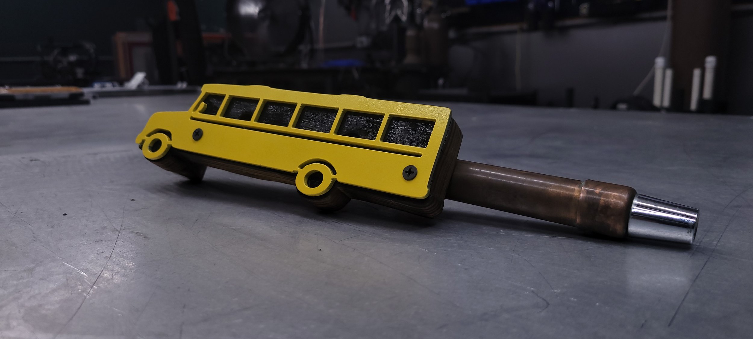 A small toy bus attached to a metal rod, resting on a flat surface in a workshop setting.