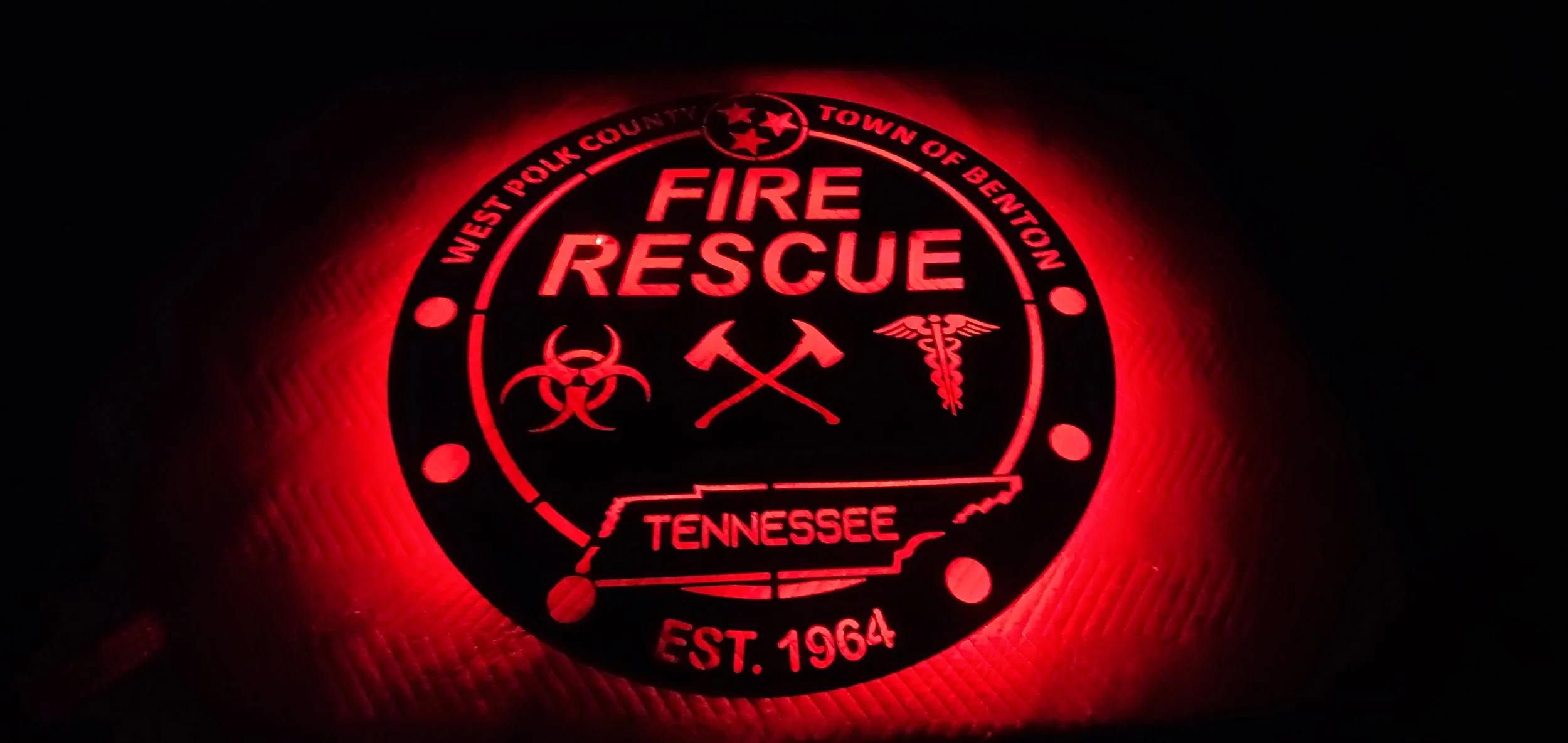 Illuminated red fire rescue badge from Tennessee, featuring symbols for biohazard, fire axe and medical caduceus, with text indicating West Polk County, Town of Benton, Tennessee, and established date 1964.