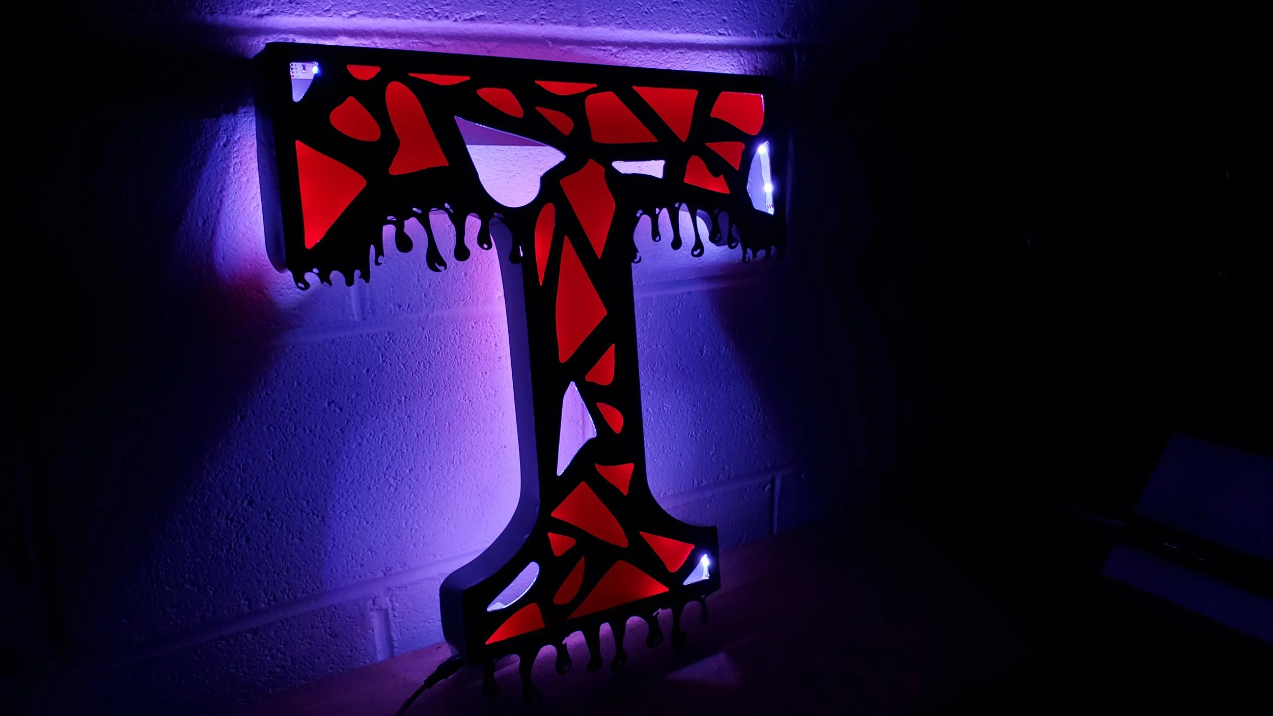 A decorative cross-shaped light fixture with stained glass panel designs, illuminated by purple and white LED lights, mounted on a wall.