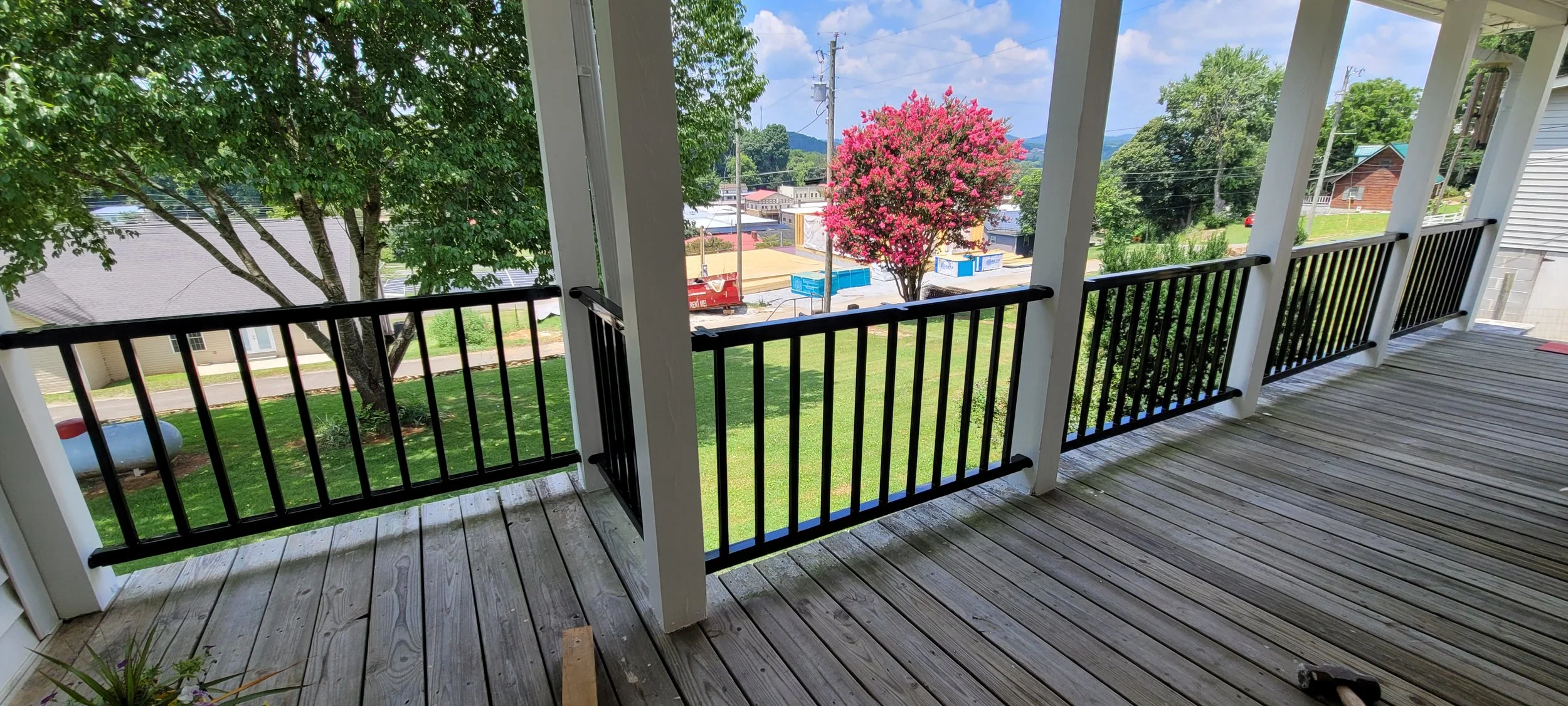 View from a covered porch with wooden flooring and black railing, overlooking a green lawn, a pink-flowered tree, and a street with buildings and utility poles.