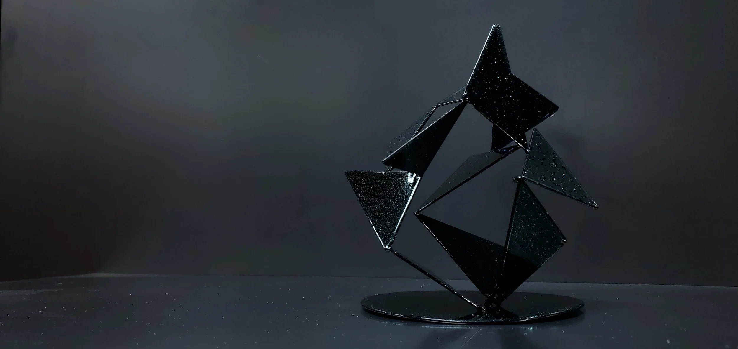 Black geometric sculpture with reflective surfaces, set against a dark background on a flat surface.