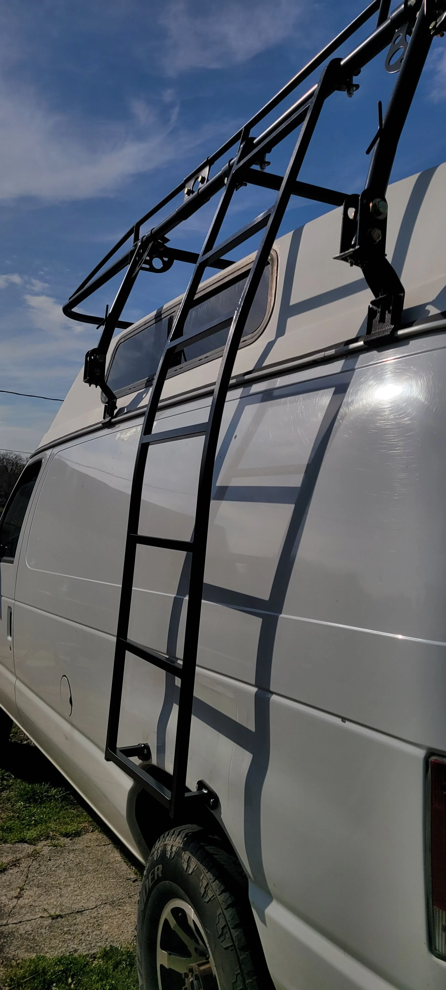 White van with a black roof rack and ladder attached to the side, parked outside under a blue sky with some clouds.