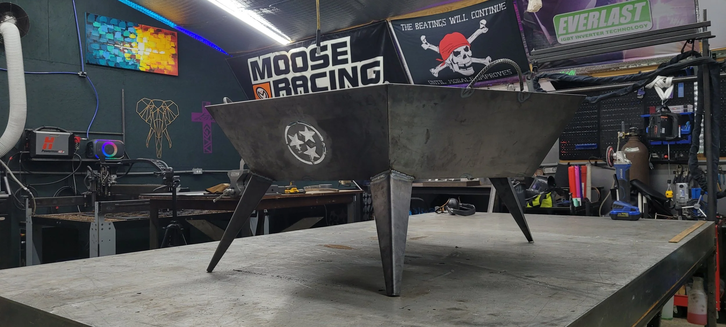 A metal fire pit with three angled legs on a work table in a workshop, with various tools, equipment, and banners in the background.