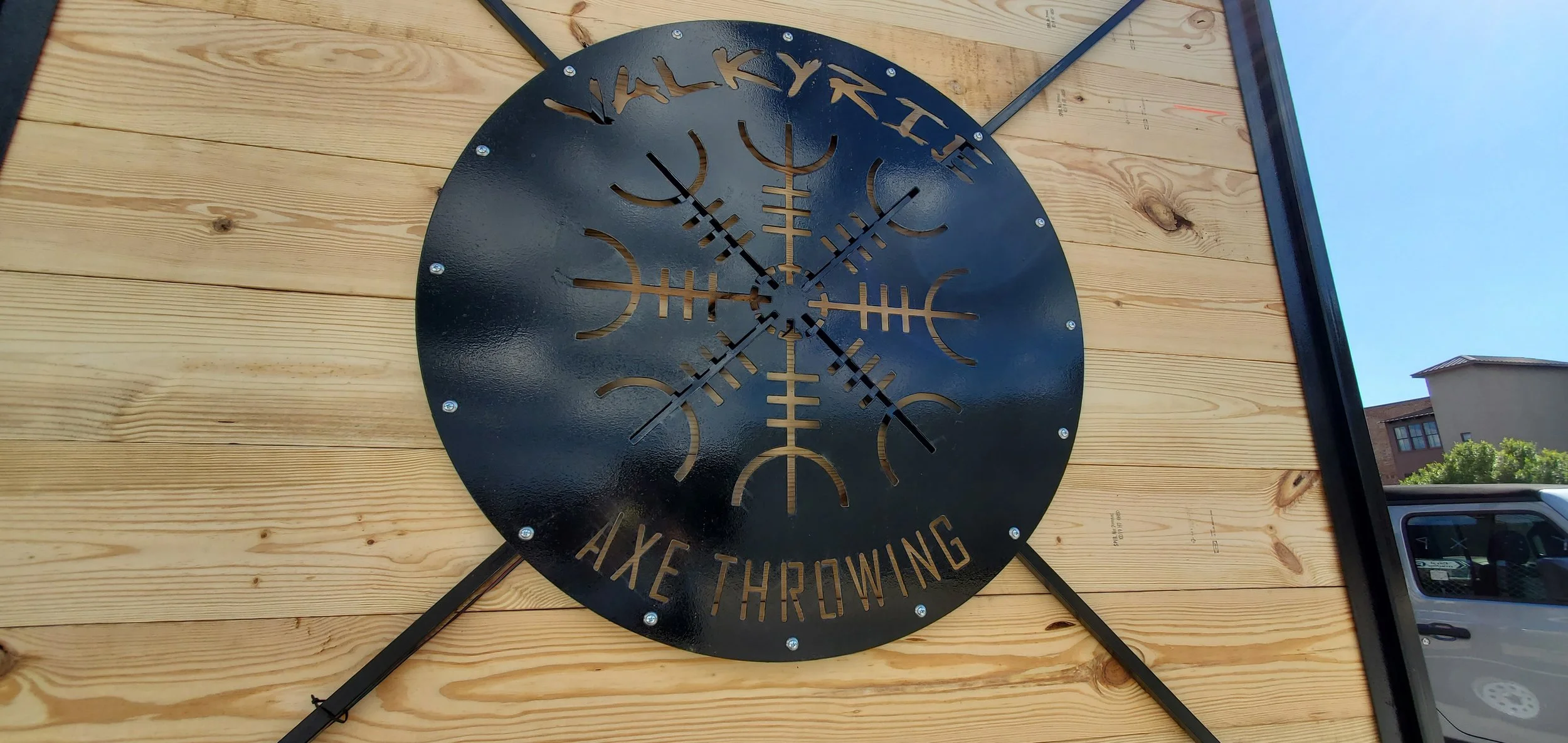 Black metal clock with Viking rune design and the words "Axe Throwing," mounted on a wooden wall outdoors.