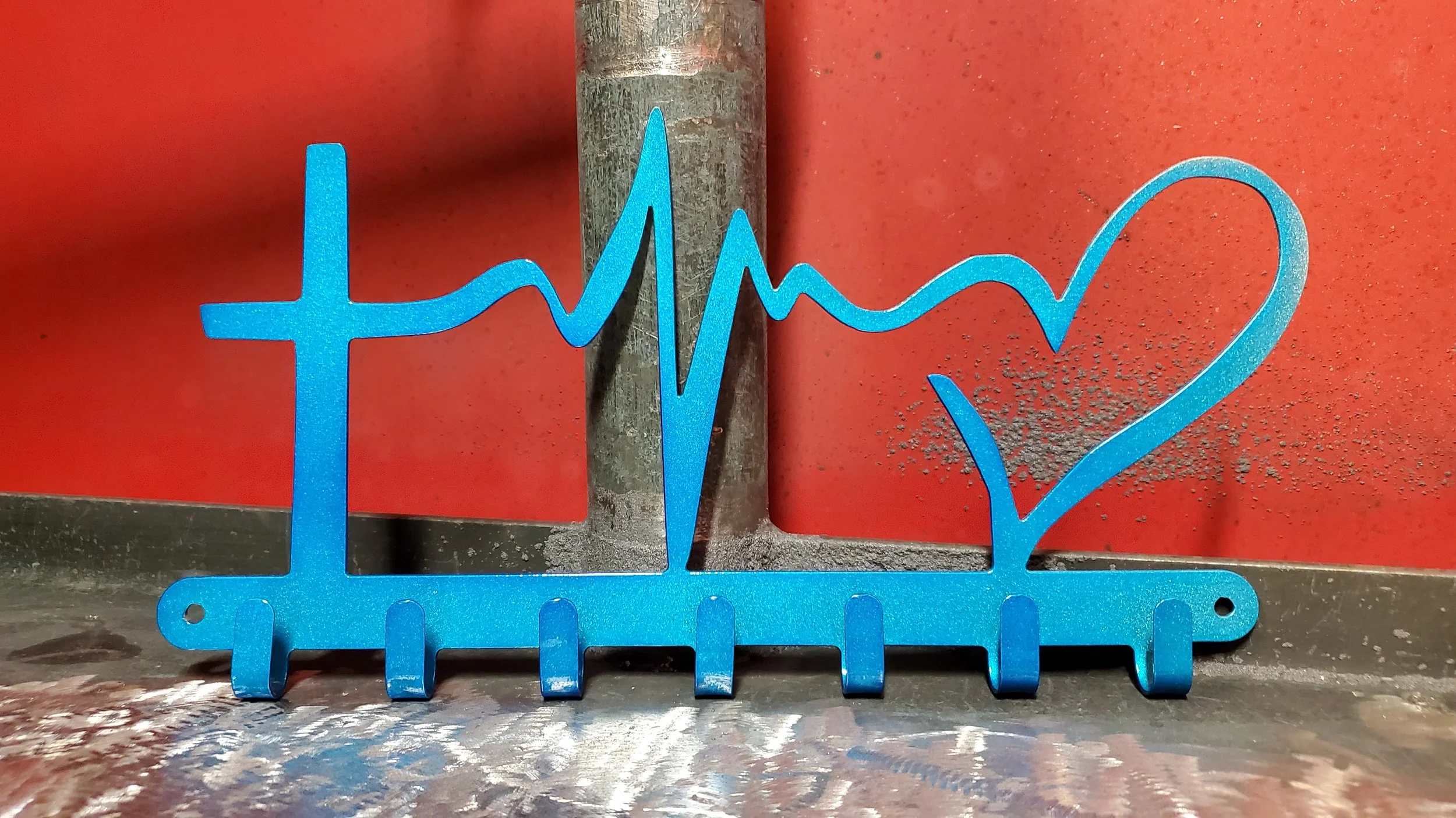 Blue metal art piece in the shape of a heartbeat and heart, with a cross on the left side, set against a red background.