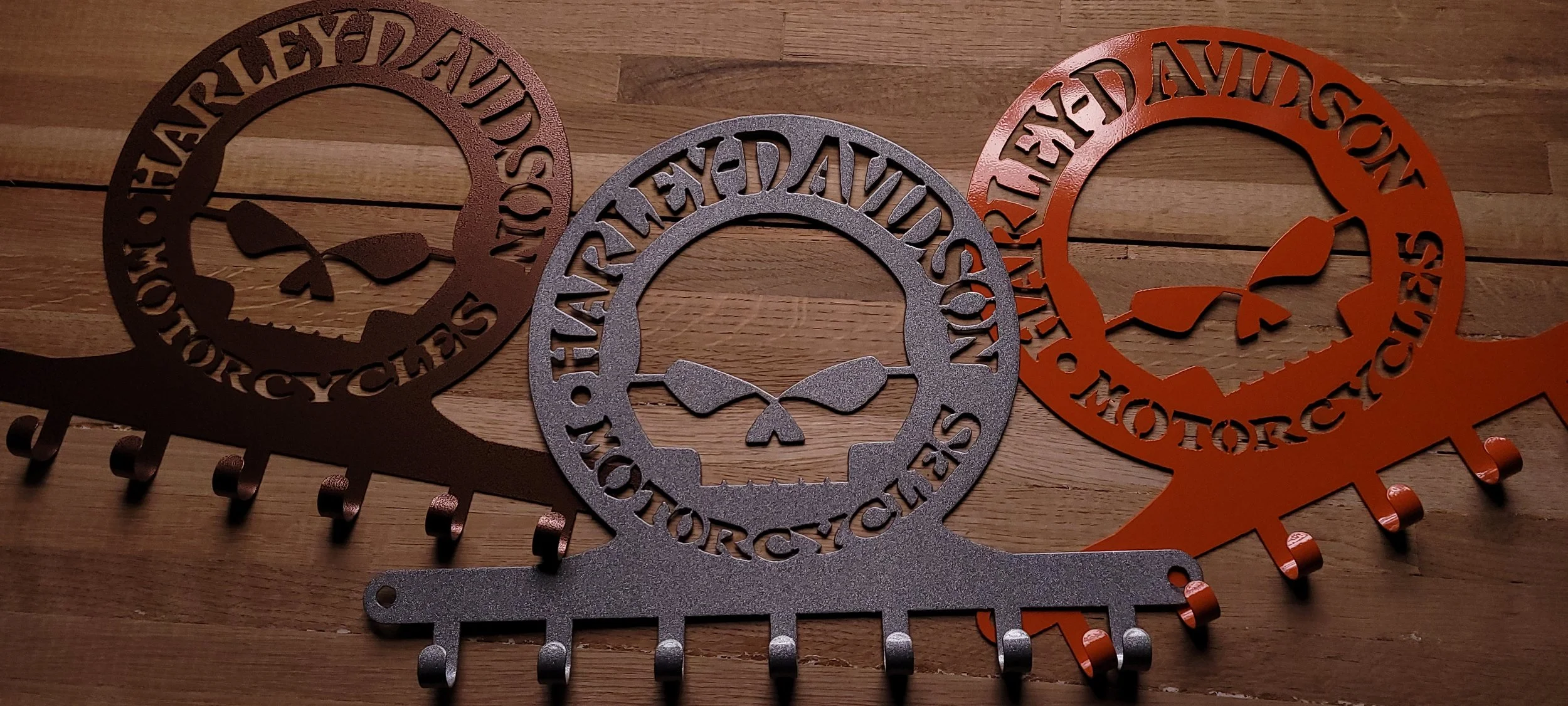 Three decorative wall hooks with Harley Davidson motorcycle logos. The hooks are circular with the words 'Harley Davidson Motorcycles' cut out around the edge, each featuring a stylized skull with motorcycle handlebars. The hooks are in three colors: