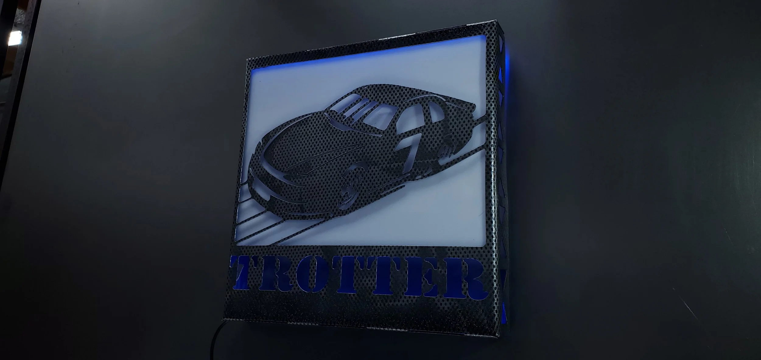 Backlit sign with a black perforated car silhouette and the word 'TROTTER' beneath it, mounted on a dark wall.