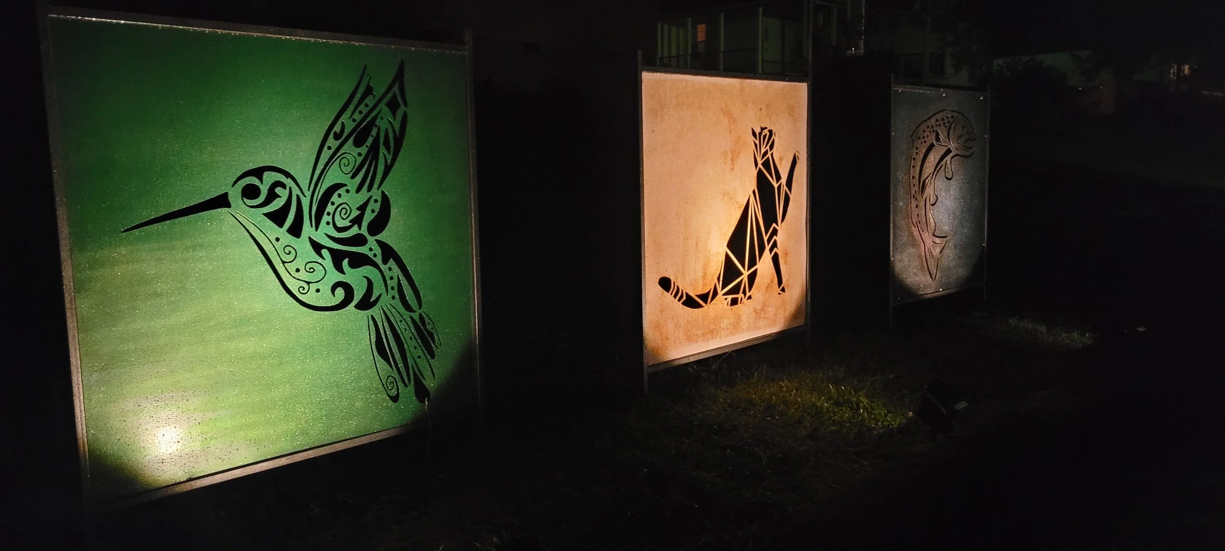 Illuminated artwork of a hummingbird, a fox, and a snake displayed outdoors at night.