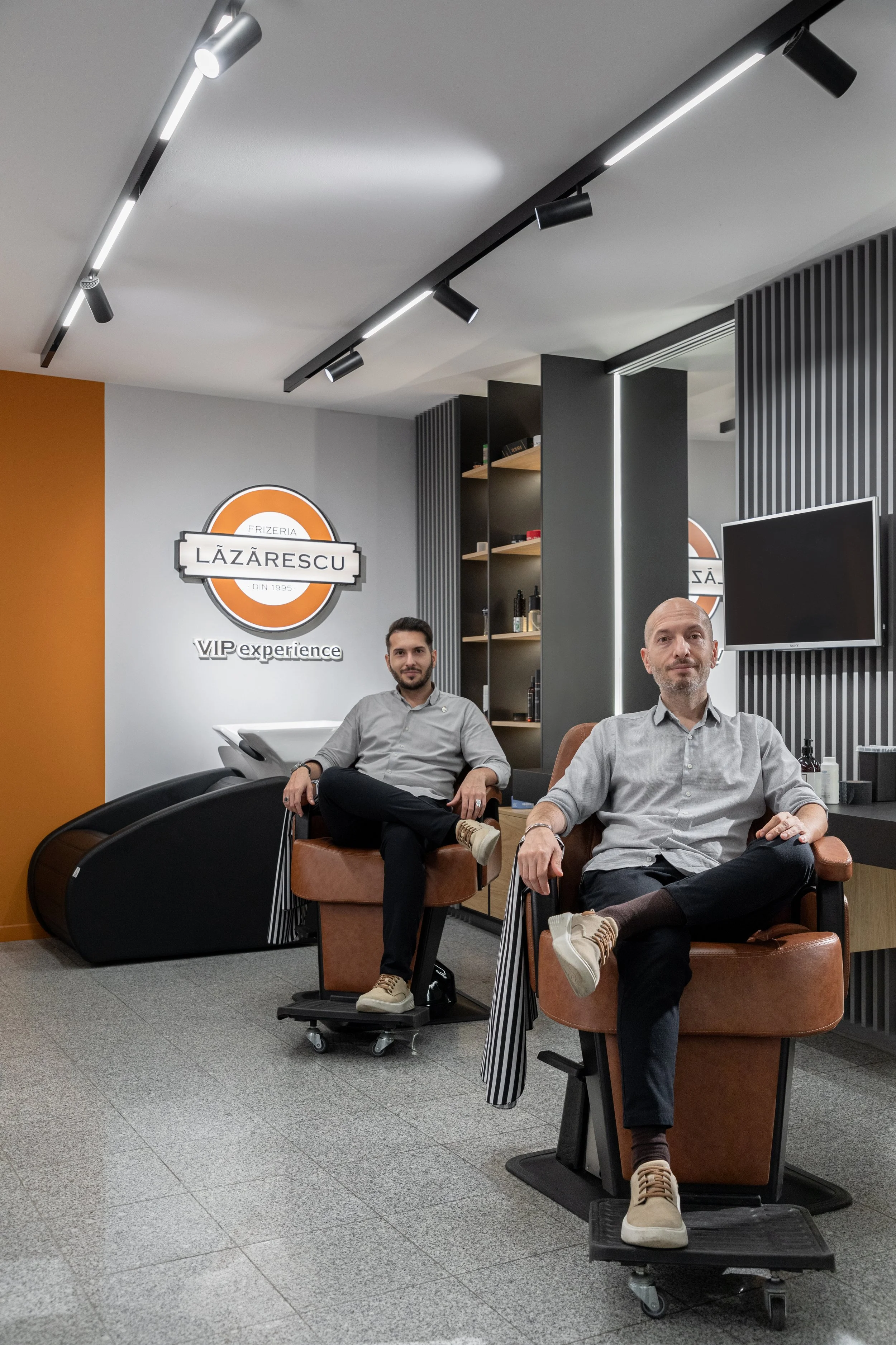 Two men sitting in chairs inside a modern salon or barbershop. The man on the left has brown hair, a beard, and is wearing a light gray shirt with black pants and beige shoes. The man on the right is bald with a gray beard, wearing a light gray shirt