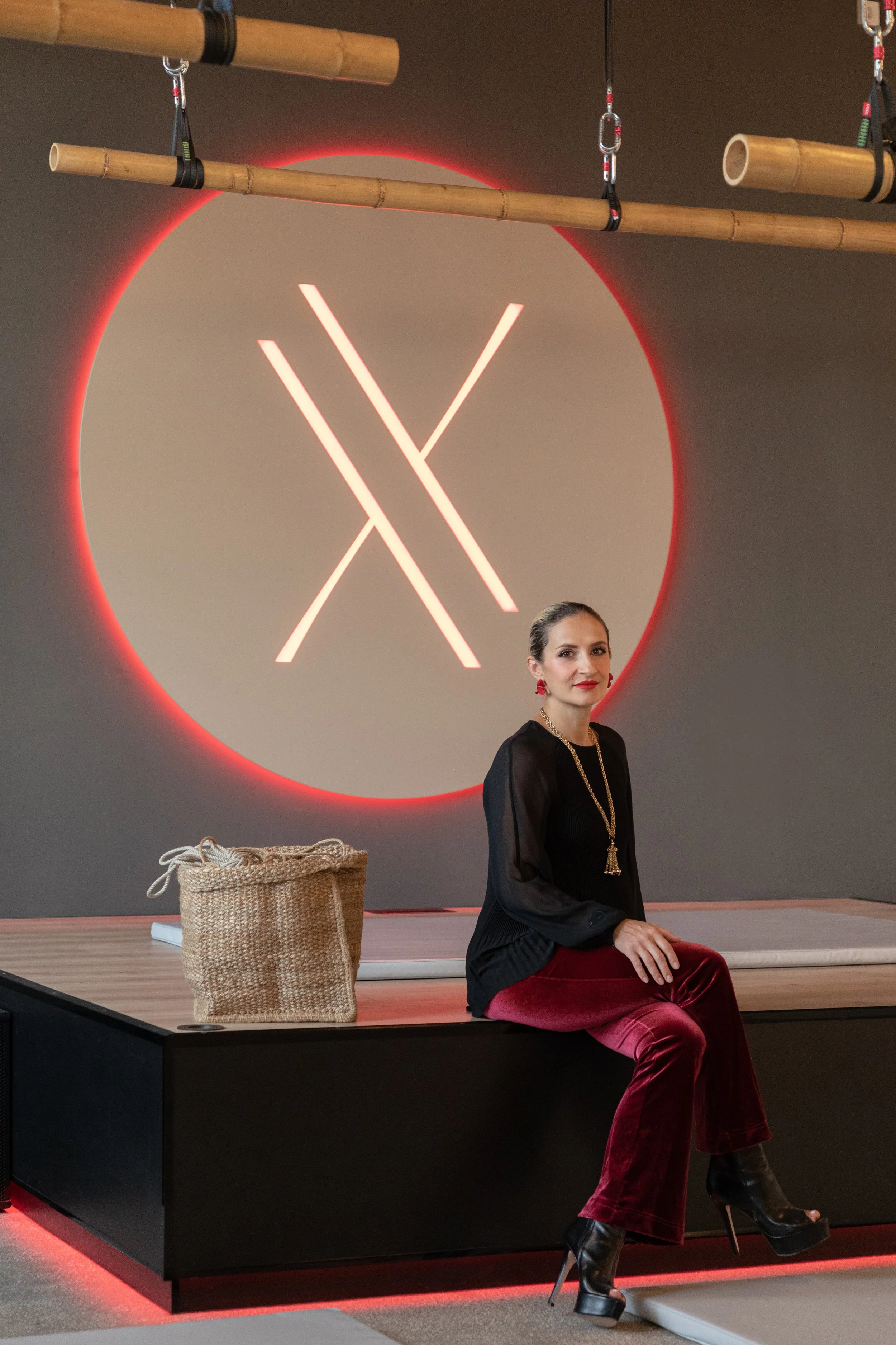 A woman dressed in black and burgundy velvet pants with high heeled boots is sitting on the edge of a platform in a yoga studio with a large illuminated logo on the wall behind her. There is a woven basket on the platform and bamboo poles hanging abo