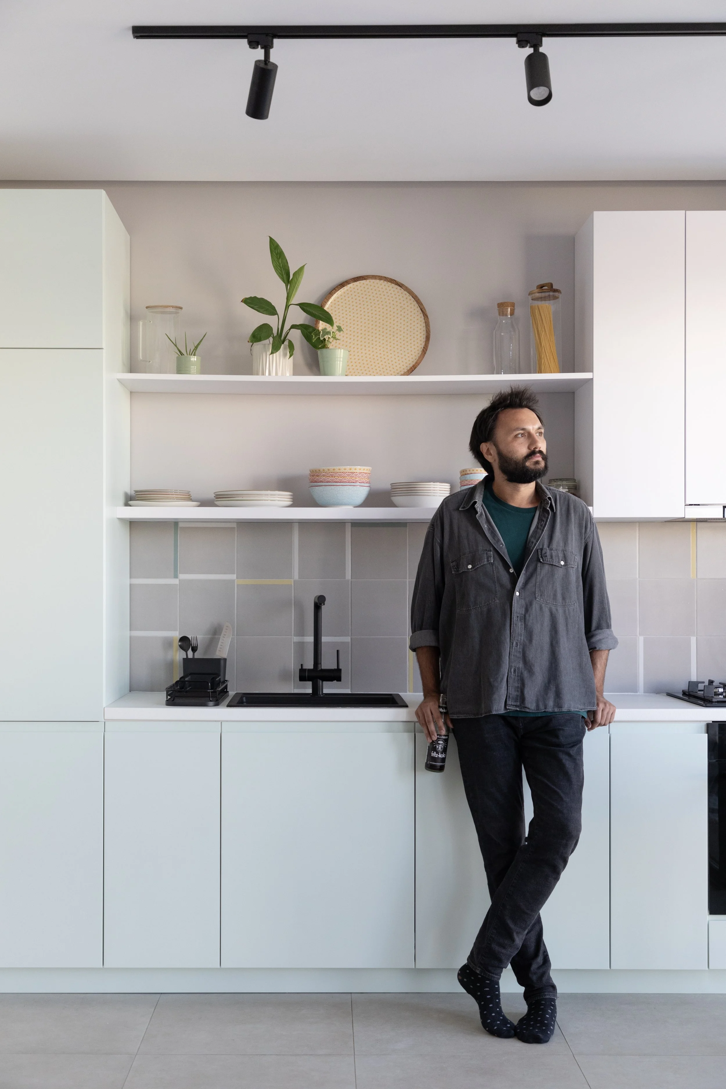 A man with a beard and disheveled hair stands in a modern white kitchen, holding a bottle of beer while leaning against the counter, wearing a black denim shirt, jeans, and polka dot socks.