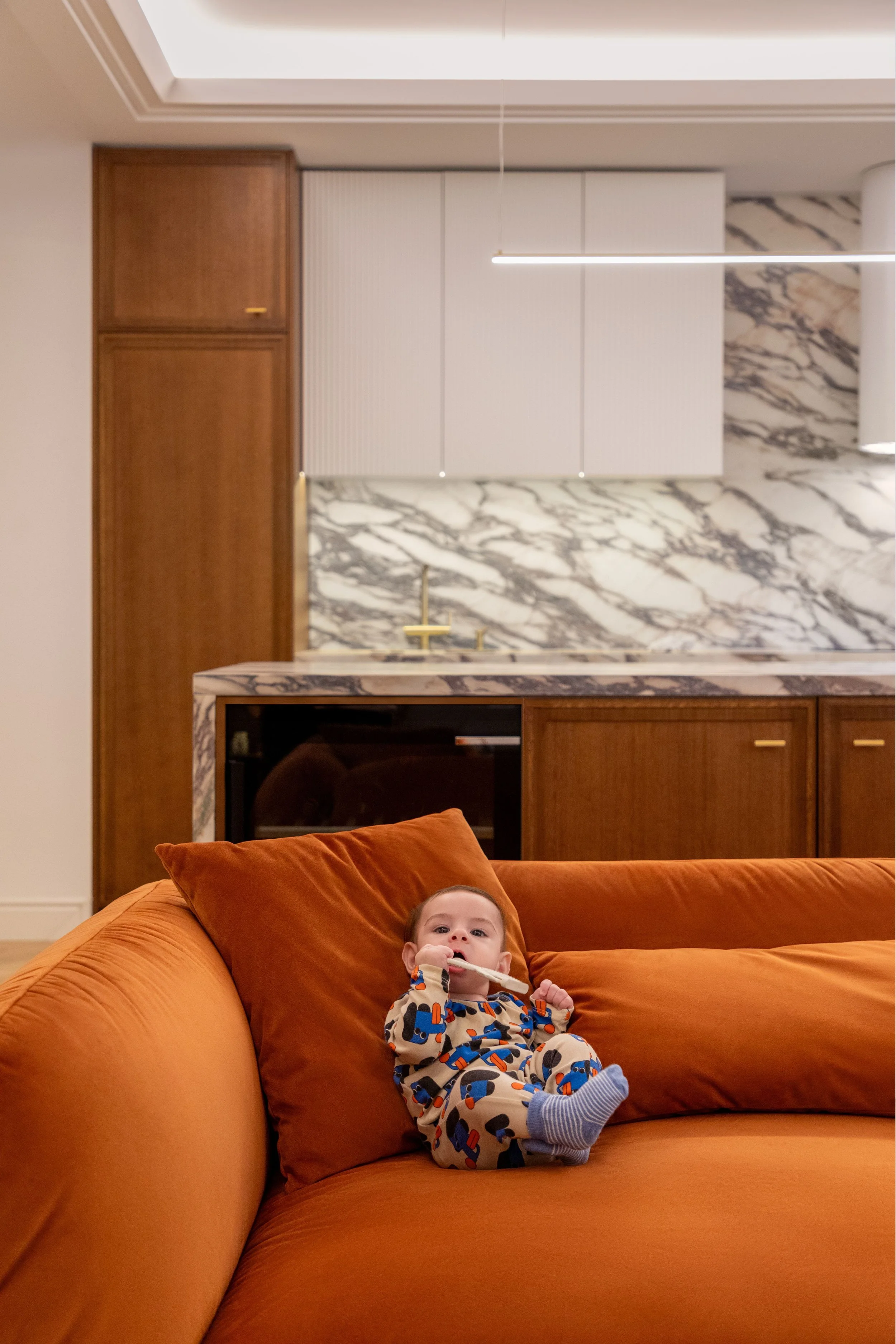 A baby dressed in colorful pajamas sitting on an orange sofa in a living room with modern decor, including a marble countertop and white cabinets, behind a large window ceiling light.