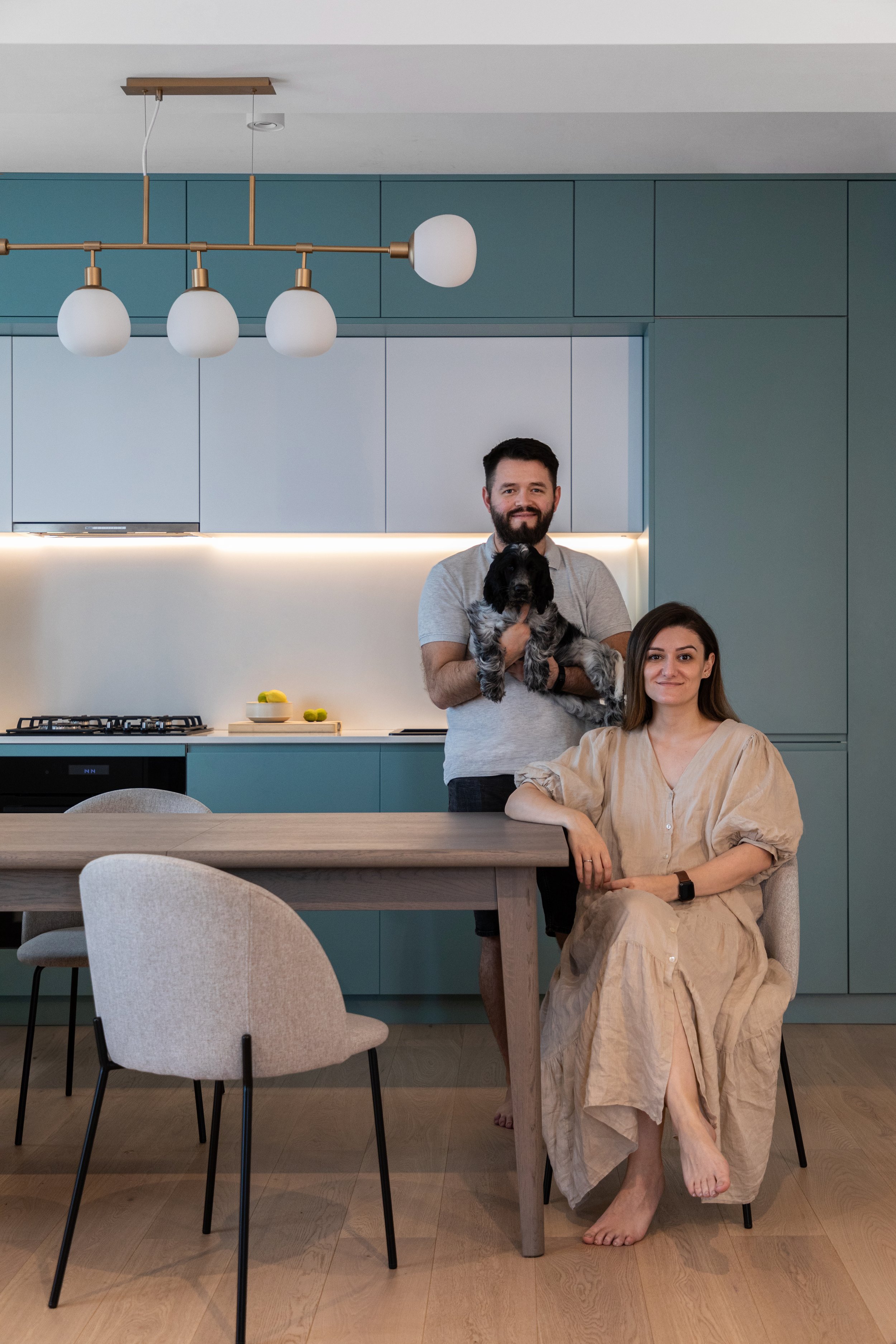 A man holding a black and white dog standing behind a woman sitting at a dining table in a modern kitchen with blue cabinets and pendant lights.