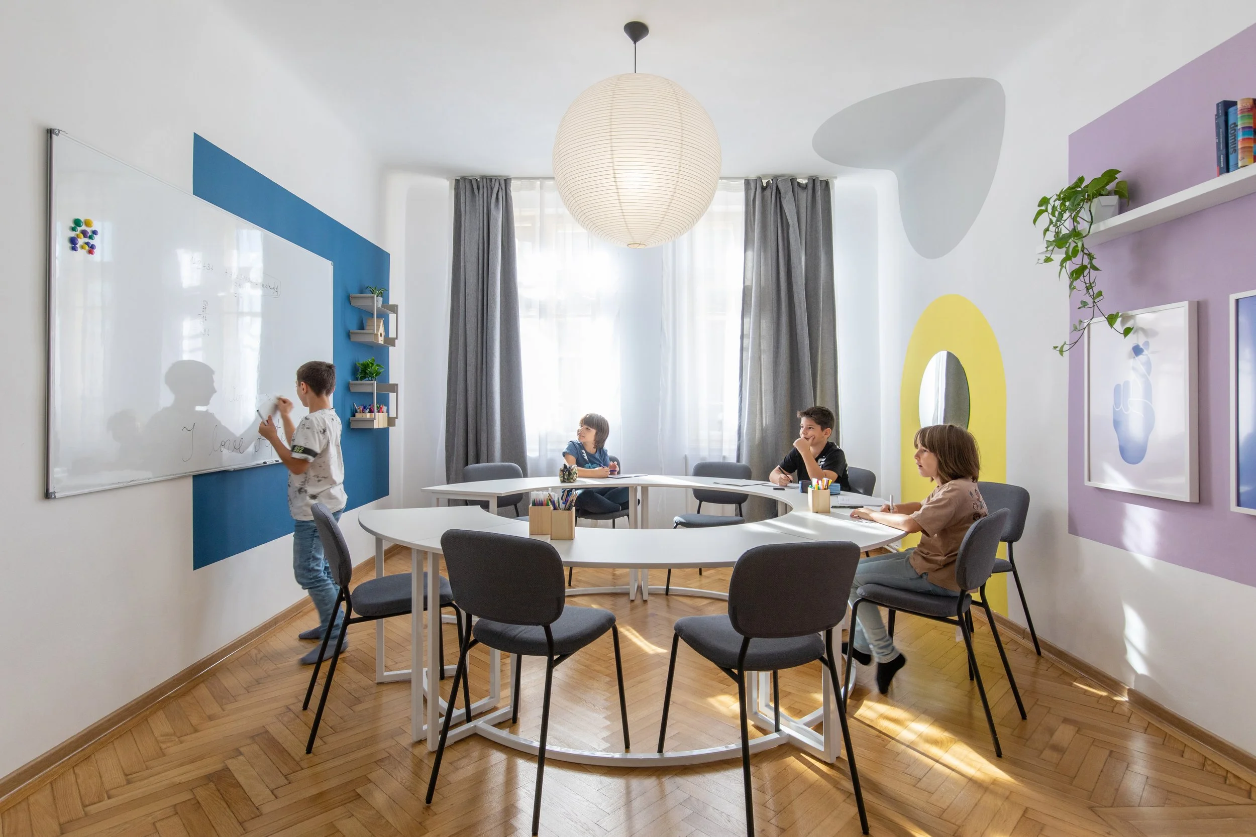 A classroom with students, a whiteboard, and colorful wall decorations. One student is writing on the whiteboard while three others sit at tables.