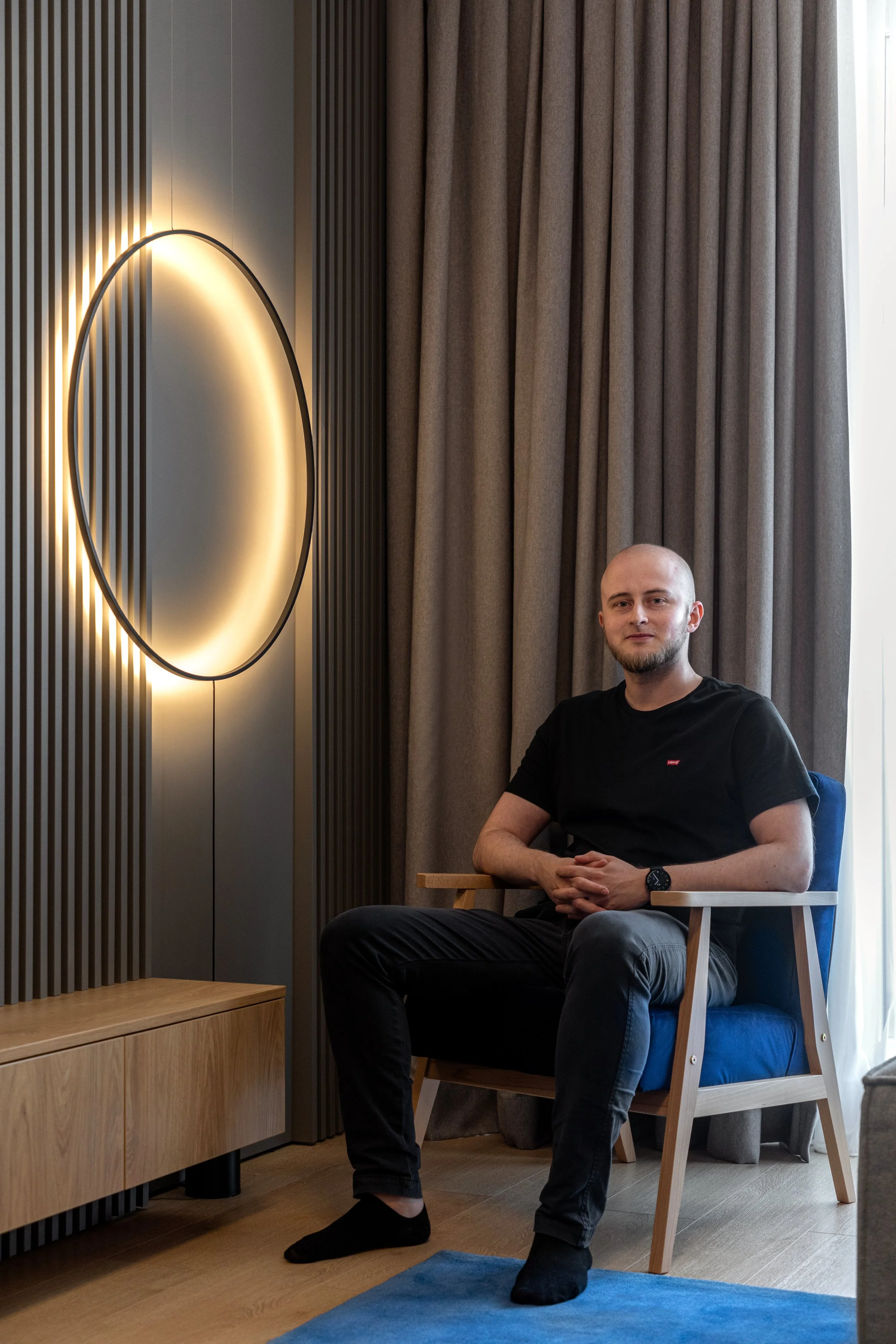 A man with a shaved head and beard sitting in a modern room with wood and gray decor, wearing a black T-shirt and gray pants, seated on a wooden chair with armrests.