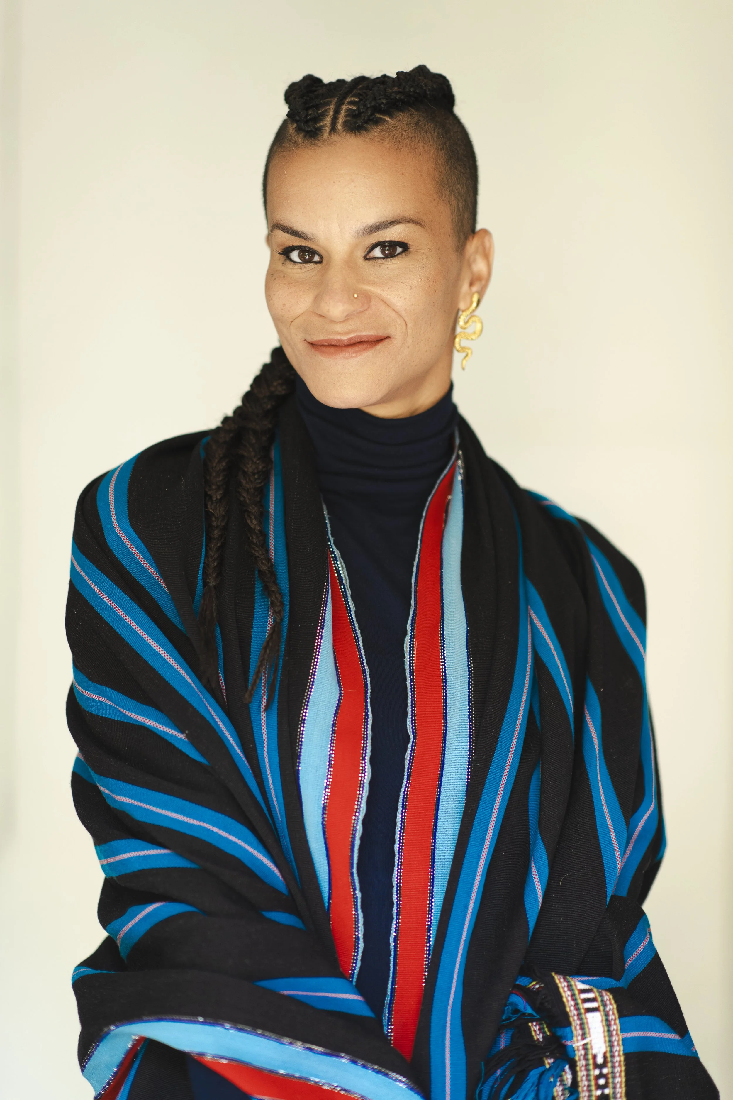 A woman with short curly hair styled in twists, wearing a black turtleneck and a colorful striped shawl with red, blue, and black stripes, gold earrings, and a nose ring, smiling against a light background.