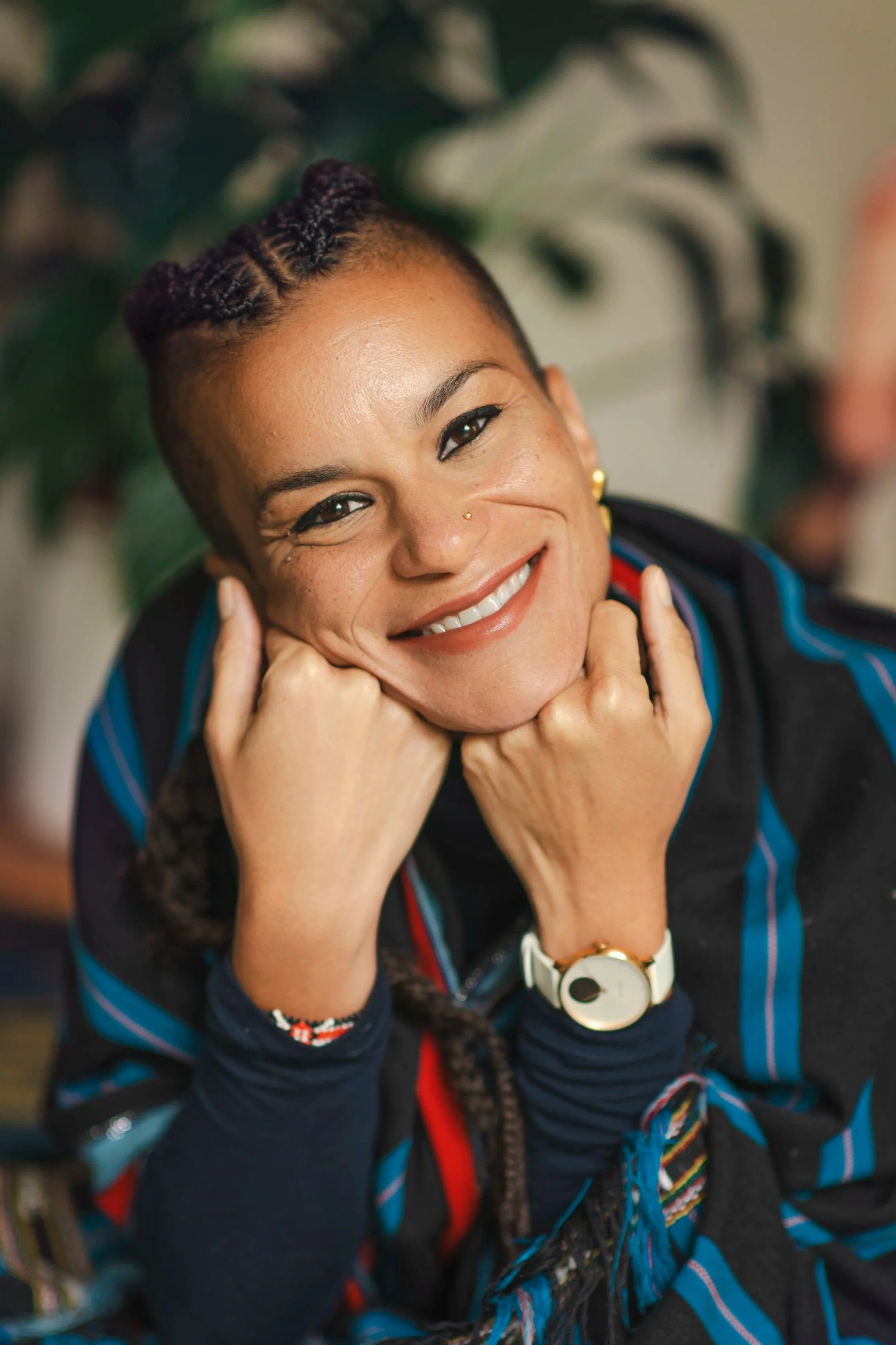 A smiling woman with braided hair, wearing a black jacket with blue accents and a white wristwatch, resting her chin on her fists.