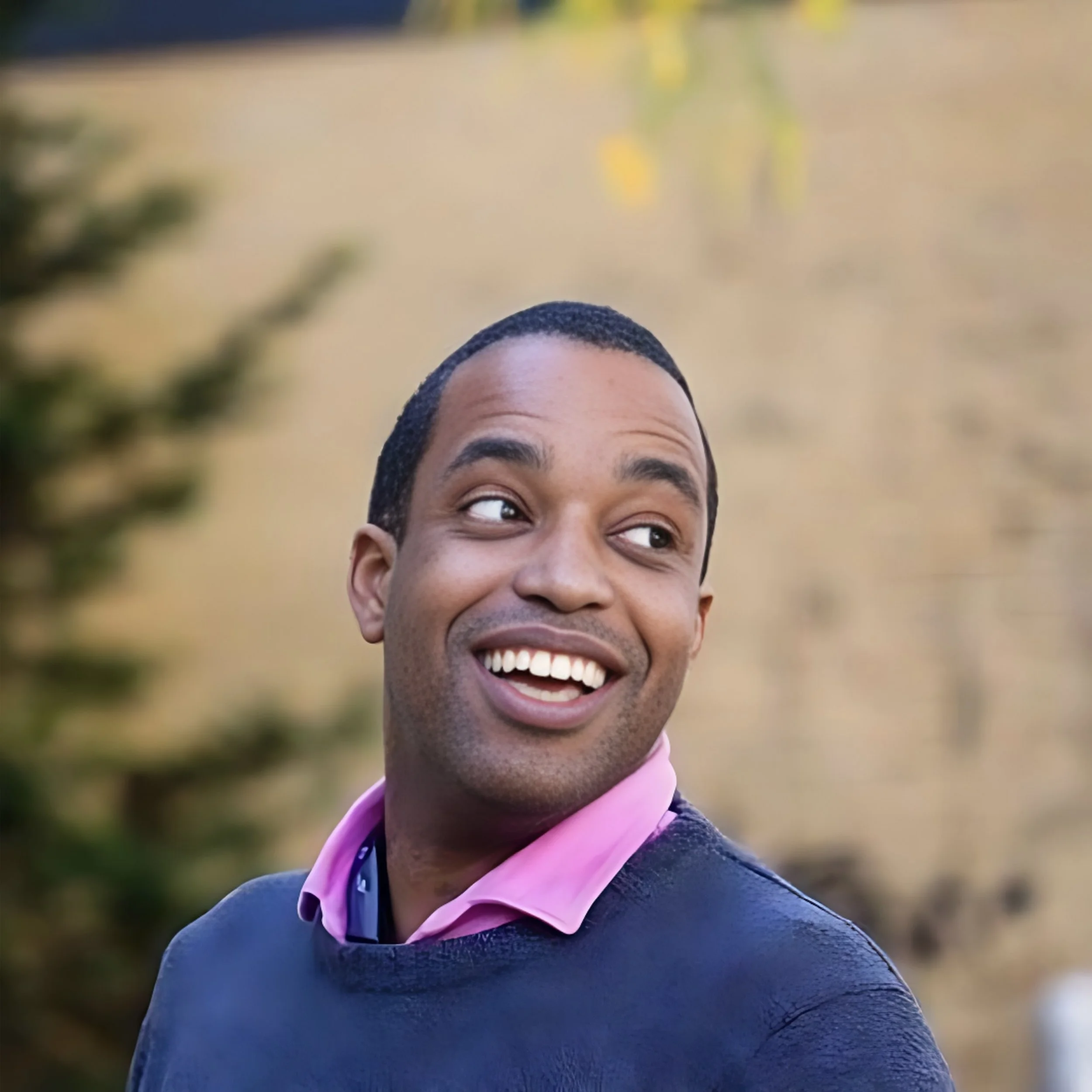 A smiling man with short dark hair wearing a pink collared shirt and a dark sweater standing outdoors with blurred trees in the background.