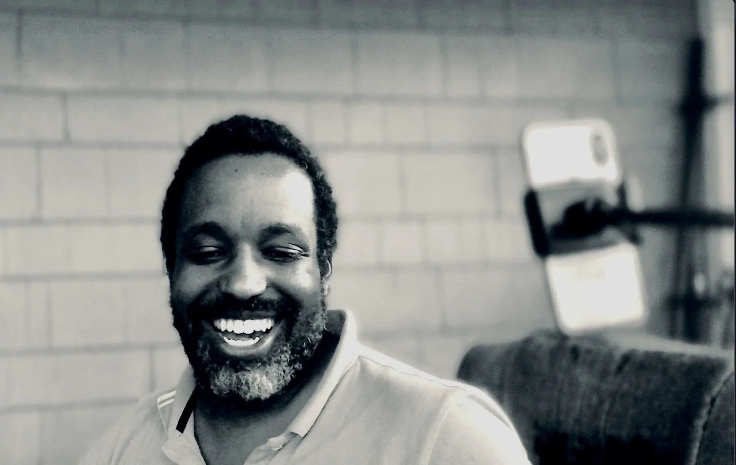 A smiling man with dark skin, curly hair, and a beard sitting in a room with a brick wall and a shelf with a phone mounted on it in the background.