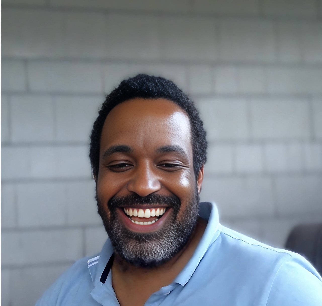 Close-up of a smiling man with dark hair, beard, and mustache, wearing a light blue shirt, against a gray brick wall.