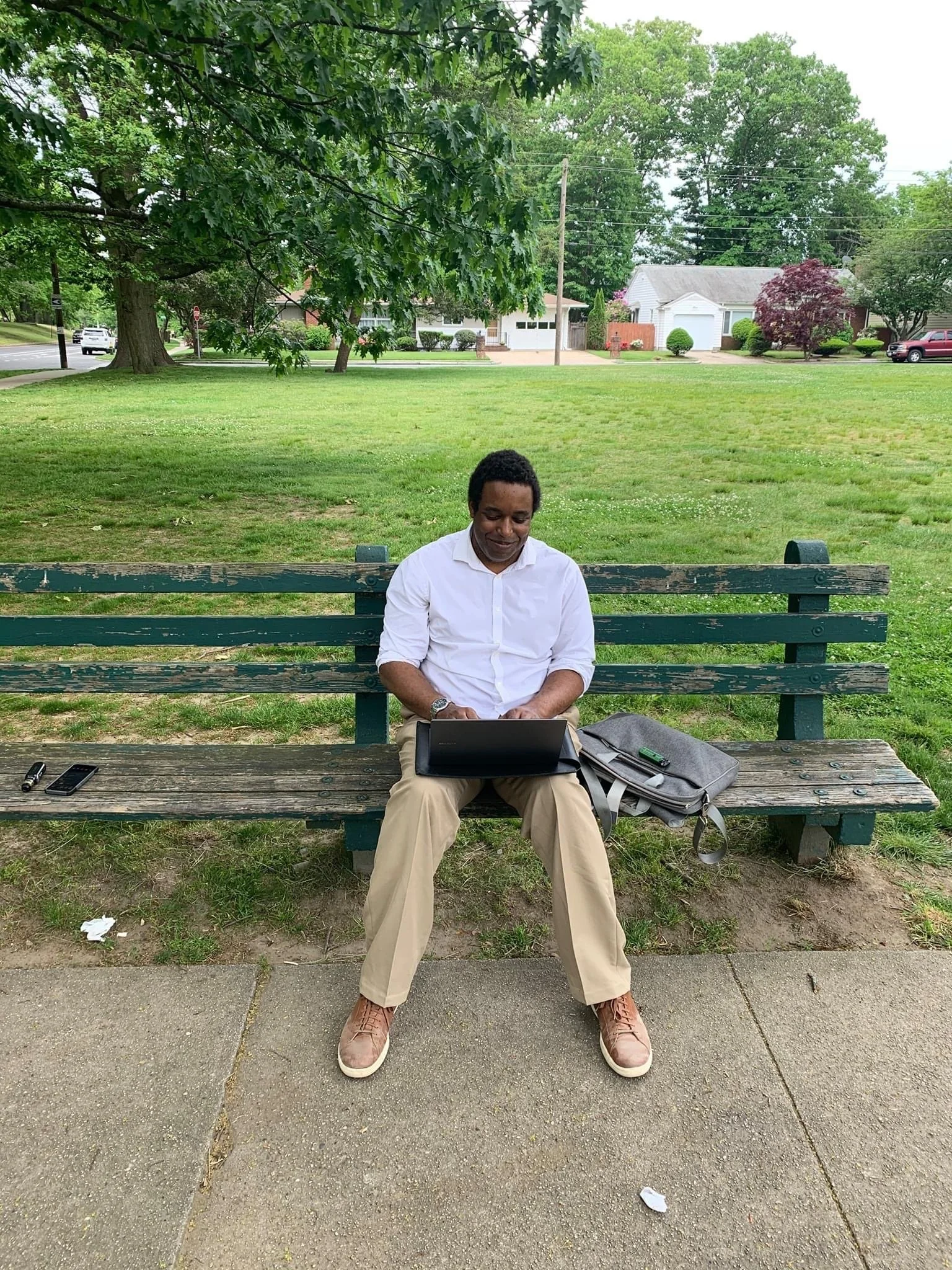 A man sitting on a park bench working on a laptop, with a backpack and two electronic devices beside him, in a green park with trees and houses in the background.
