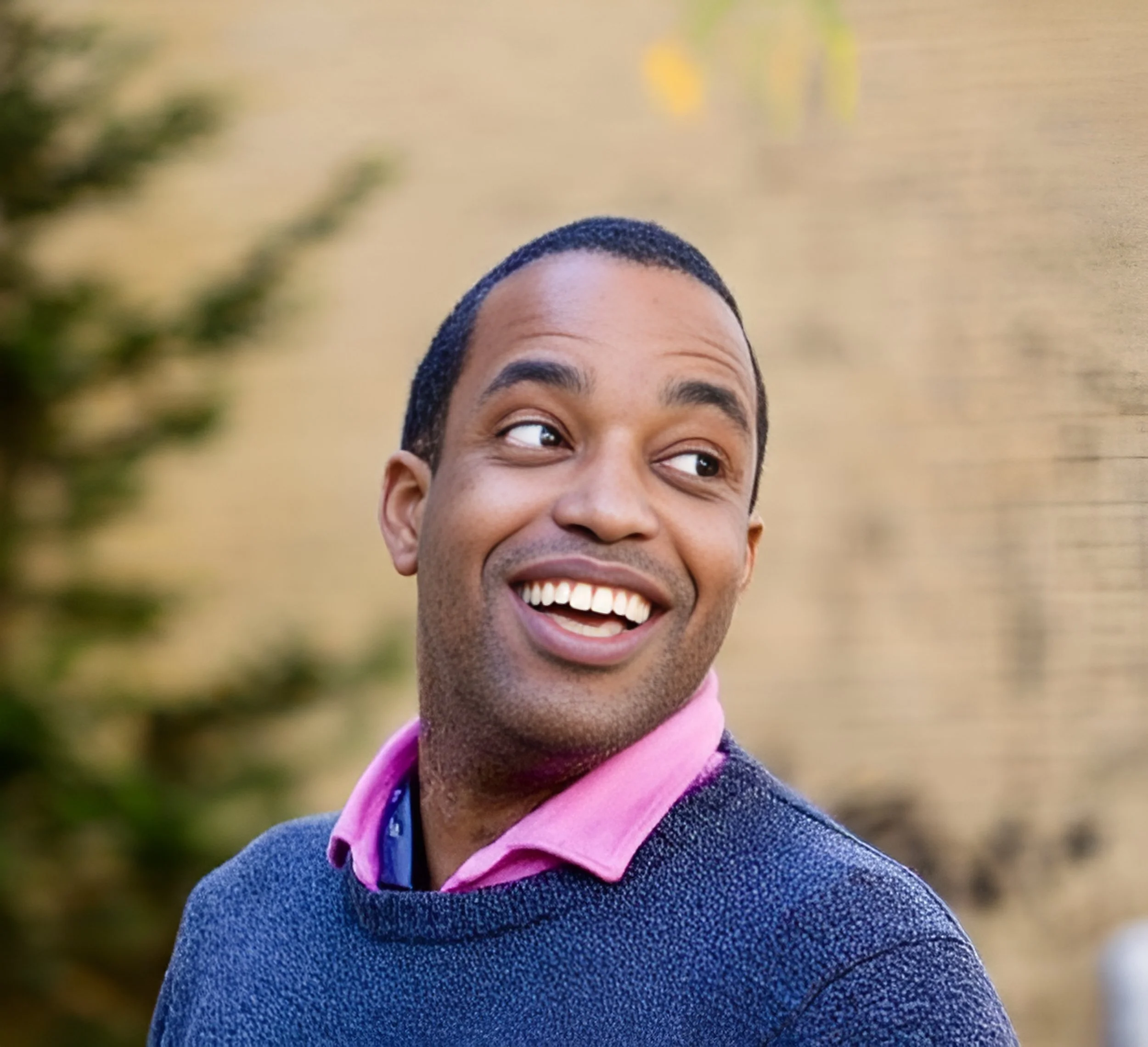A man smiling and looking to the side outdoors with a blurred yellowish background.