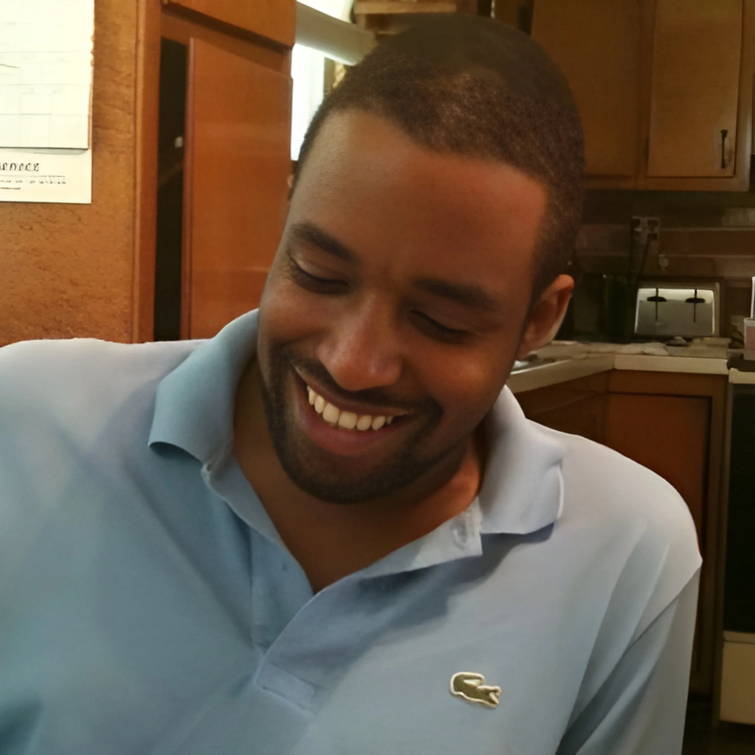 A smiling man with short hair and a beard wearing a light blue polo shirt indoors in a kitchen.