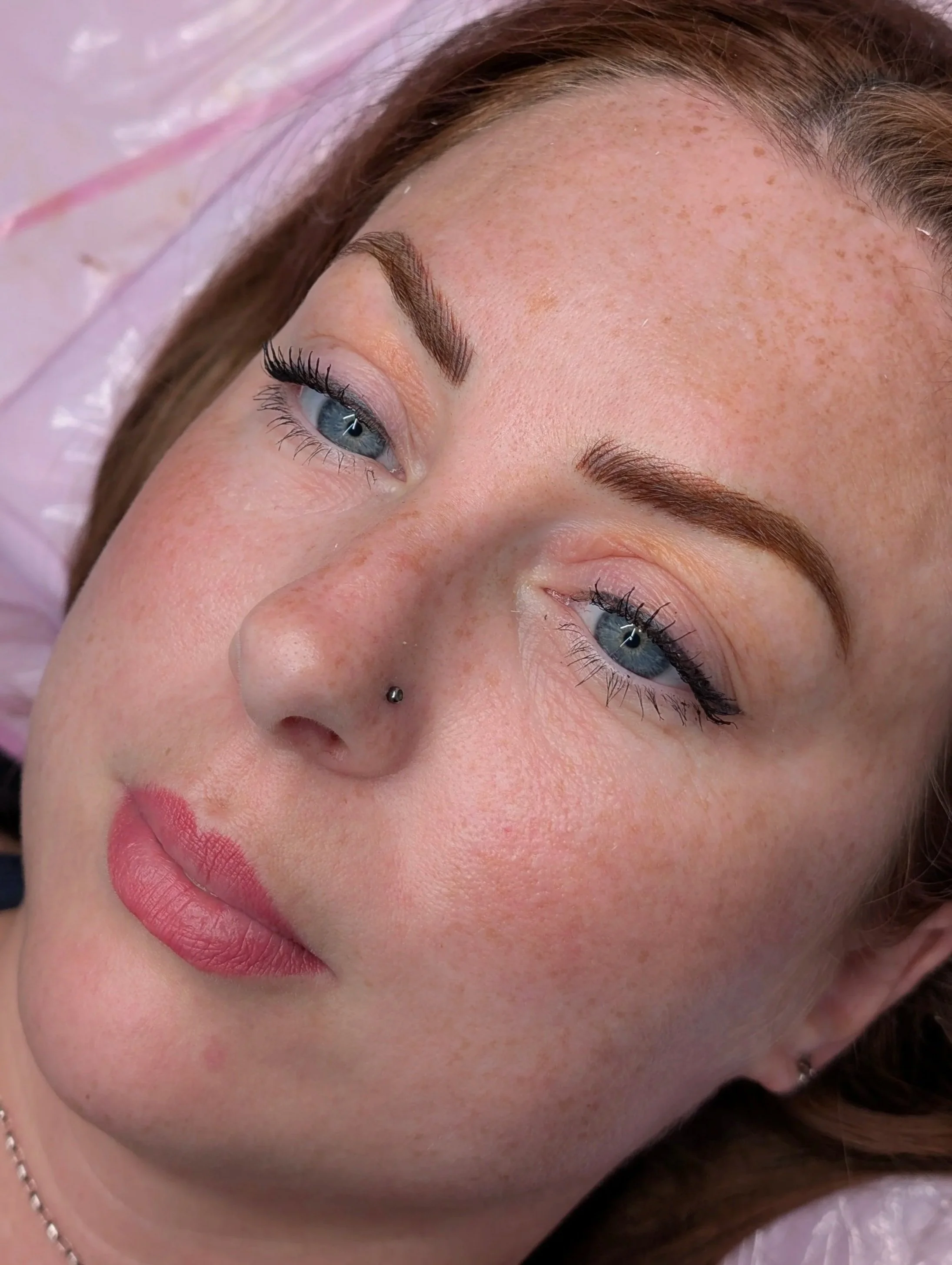 Close-up of a woman's face with makeup, freckles, blue eyes, and a piercing on her nose.