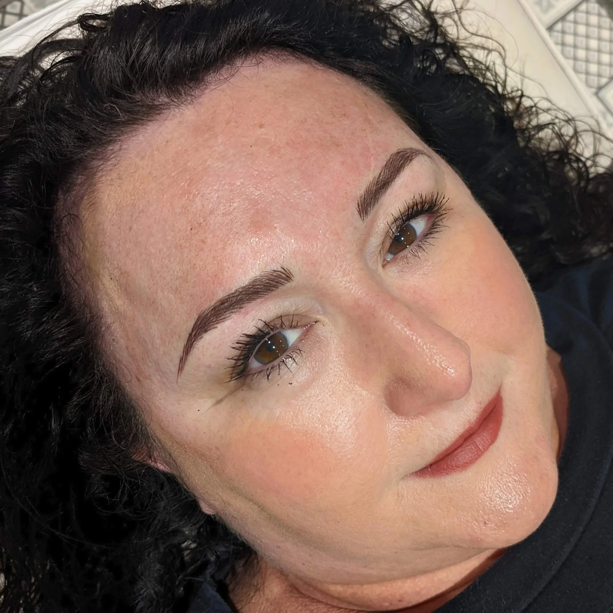 Close-up of a woman's face showing her forehead, eyebrows, and part of her eye with long eyelashes, and her curly black hair. She has light skin and appears to be lying down.