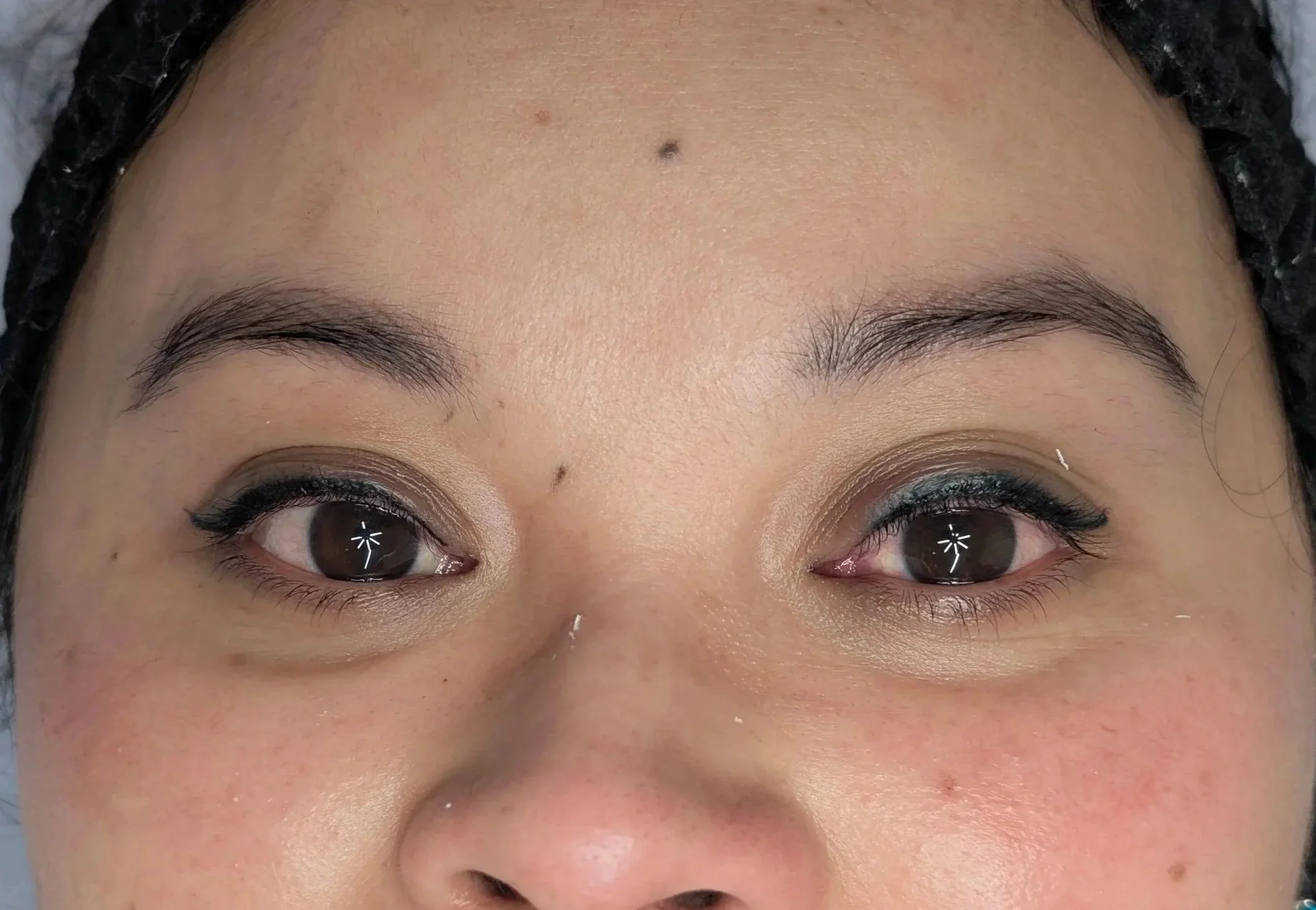 Close-up of a woman's face, showing her forehead, eyebrows, and eyes, with smooth skin and dark hair partially covered by a black hair covering.