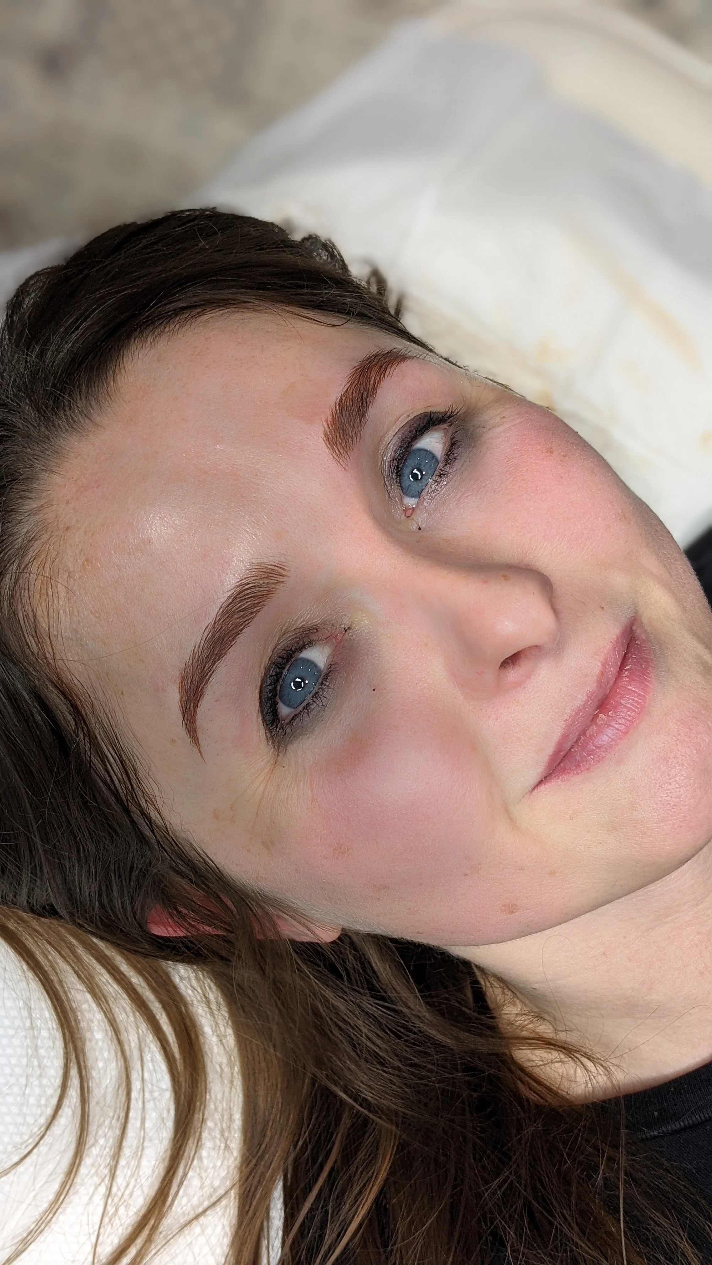 Close-up of a woman's face lying on a surface with her eyes open, showing her facial features, makeup, and brown hair.