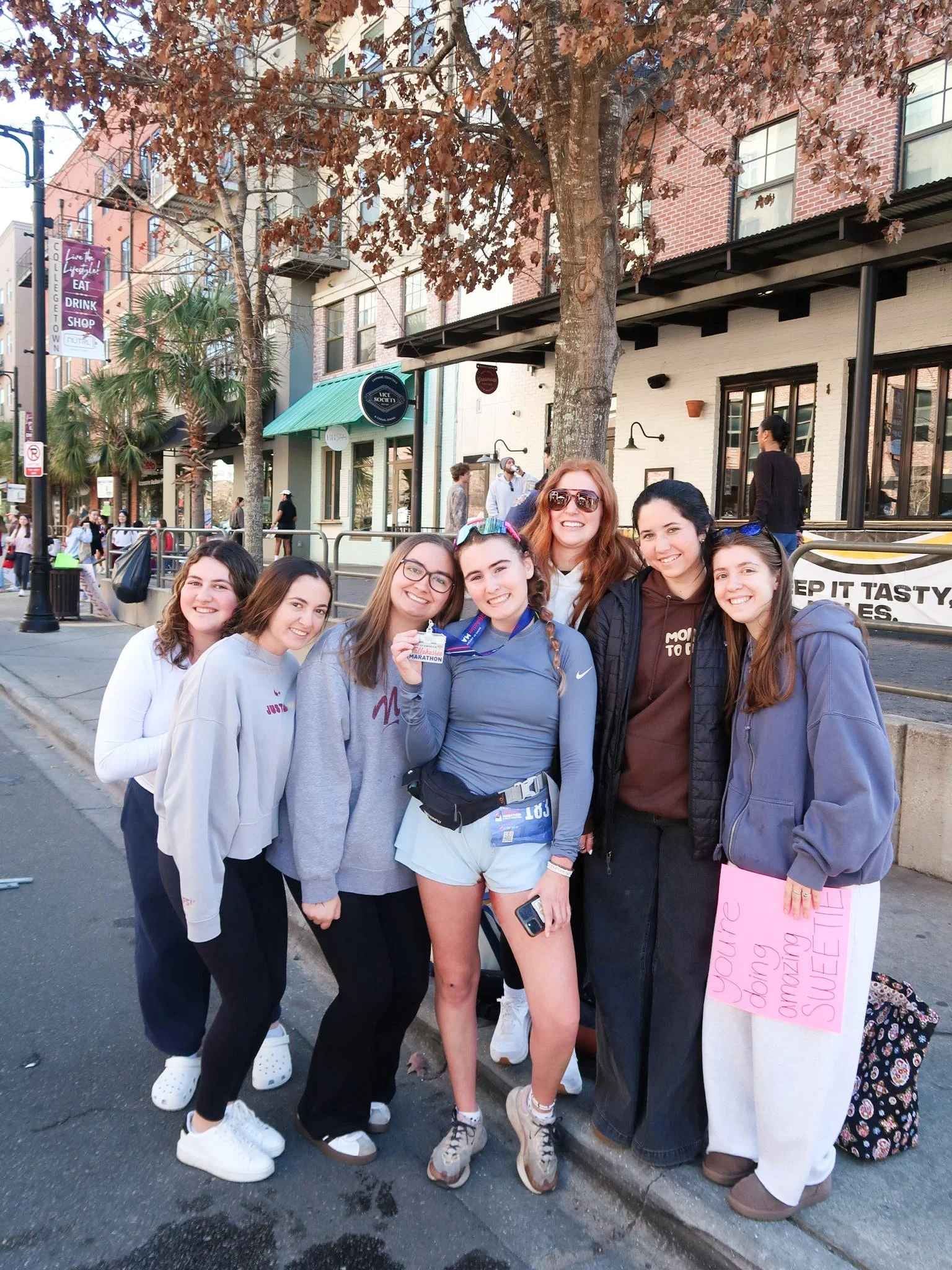 Group of eight women standing on a sidewalk, smiling at the camera, with trees and buildings in the background. One woman is holding a marathon medal, and another is holding a pink sign that says 'You're doing amazing sweetie.'