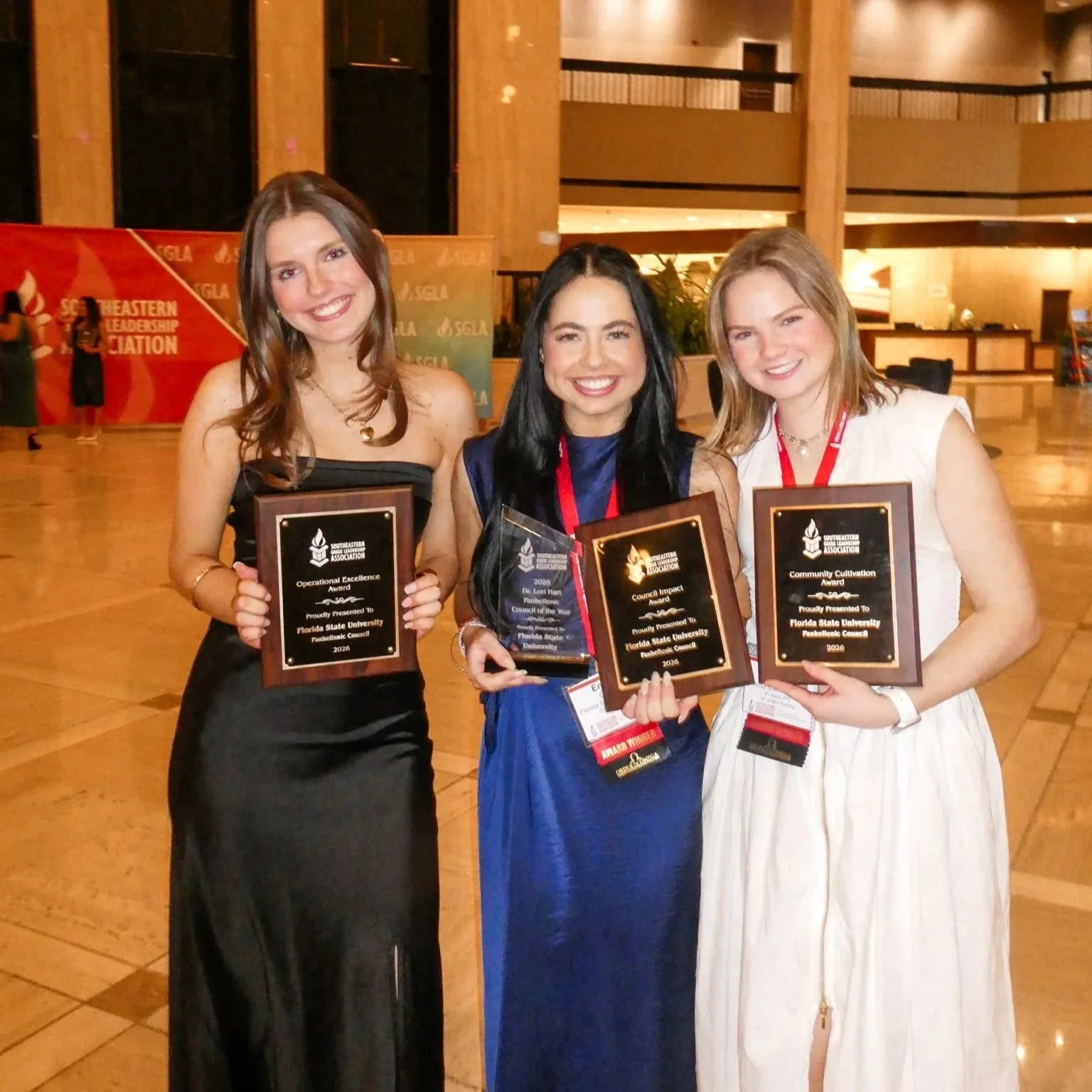 Three women in formal attire holding awards at an event in a large indoor venue with wood paneling and a banner in the background.