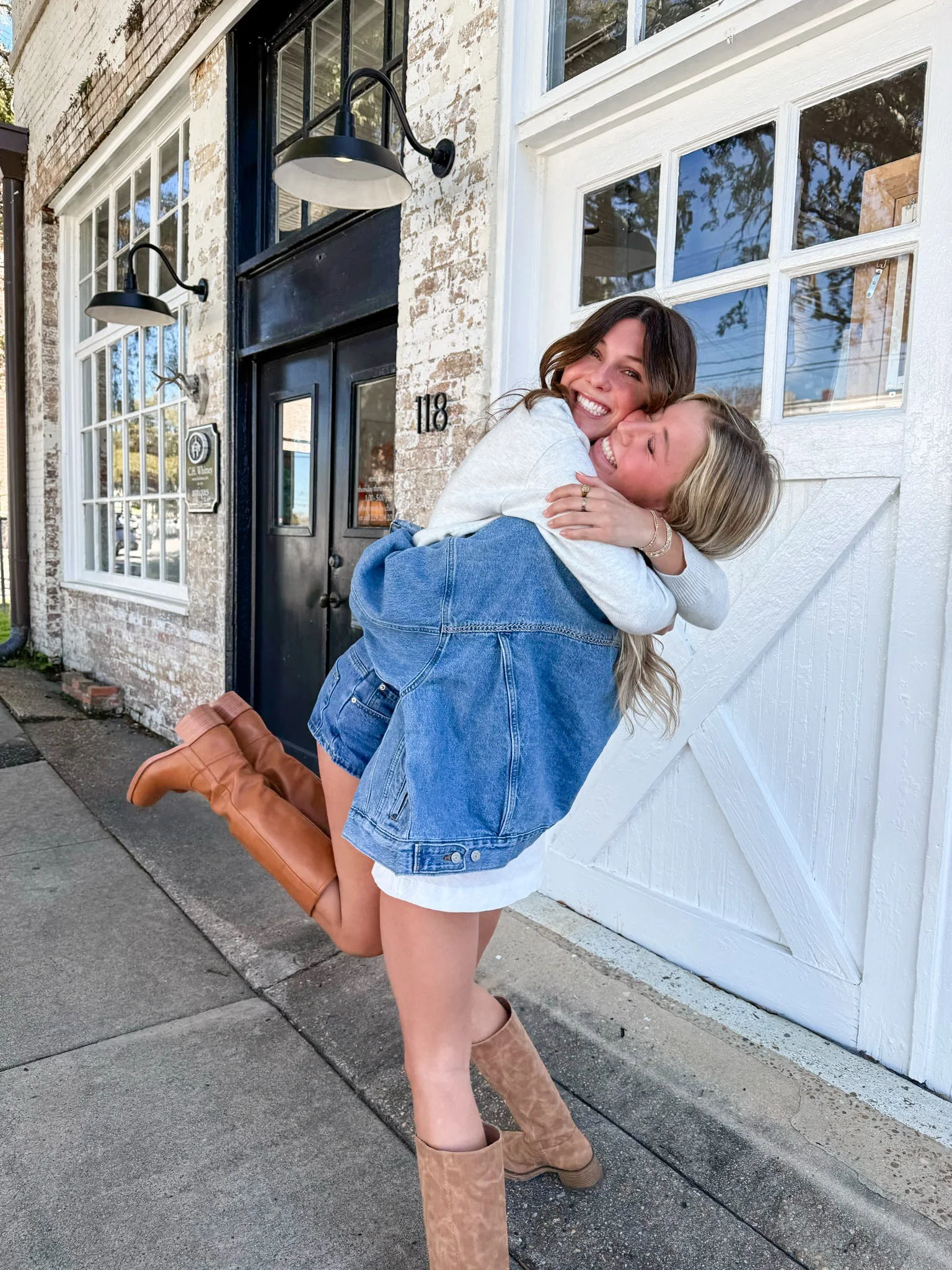 Two young women smiling and hugging each other, with one lifting the other off the ground in front of a brick building with large windows and a white door.